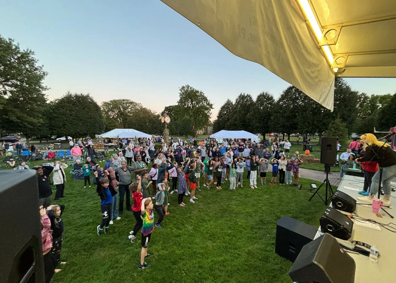 People gathered in a park at dusk, participating in an outdoor event with a stage where a band is performing. Some attendees are dancing, and there are tents and trees in the background.
