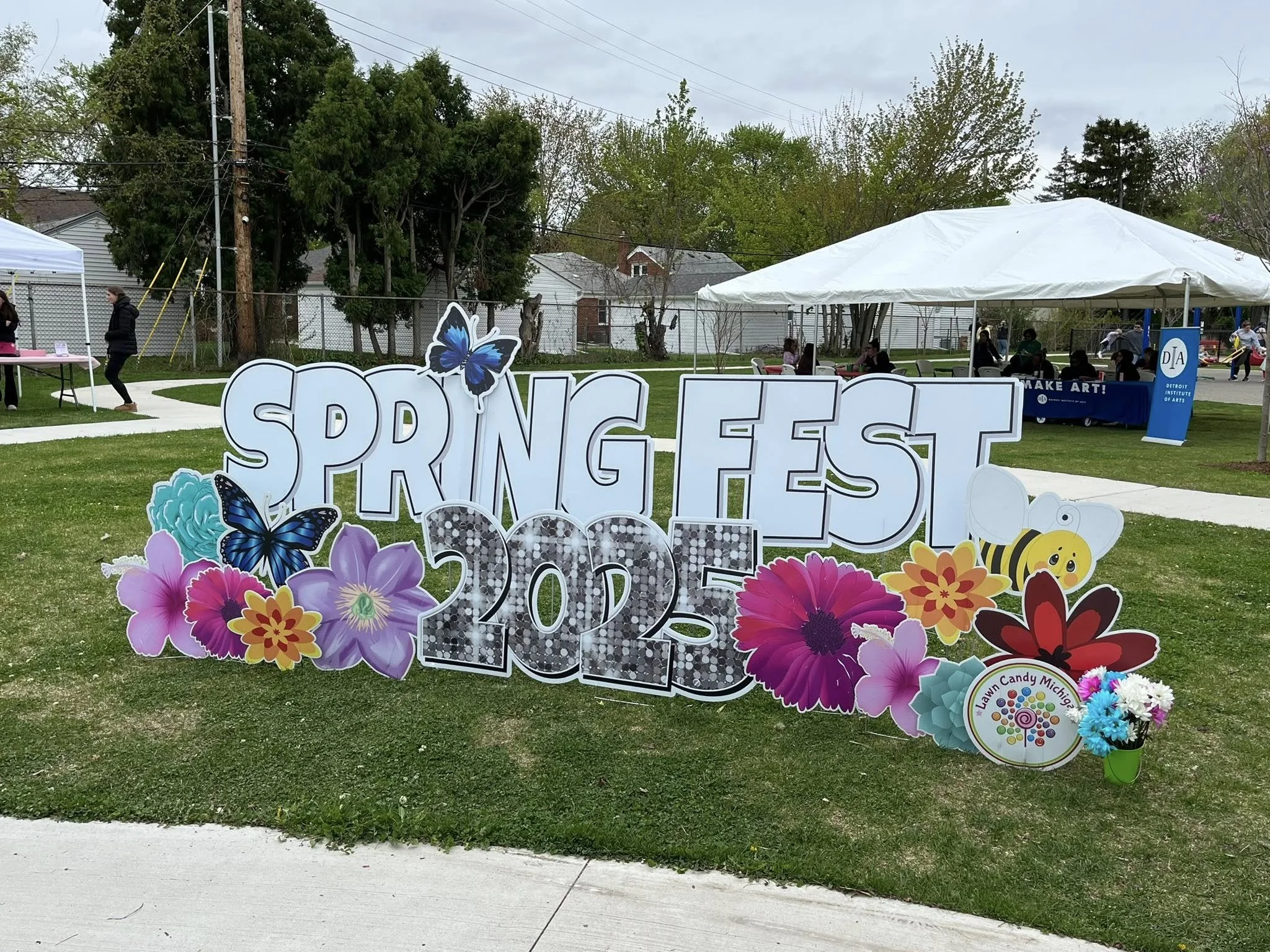Decorative sign reading 'Spring Fest 2023' with colorful flowers and butterflies at an outdoor event, with tents and people in the background.