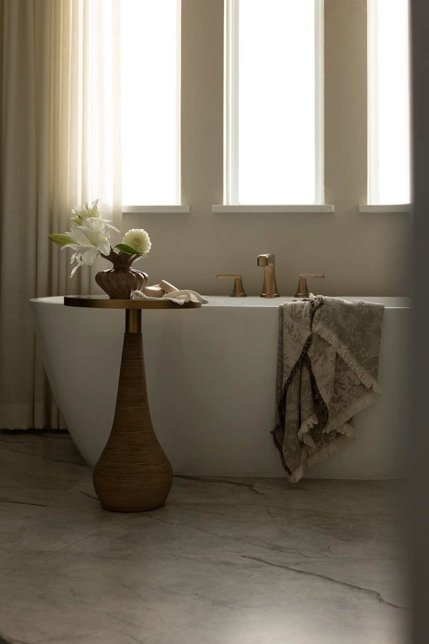 An elegant white bathtub with  soft light filtering in from the large window in the background. An accent table is in the front of the bathtub holding elegant bath accessories. 