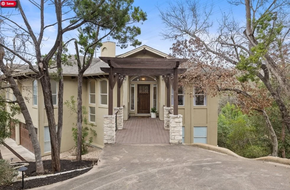 A house with a covered front porch, stone pillars, wooden beams, and a dark front door, surrounded by trees and a sloped driveway.