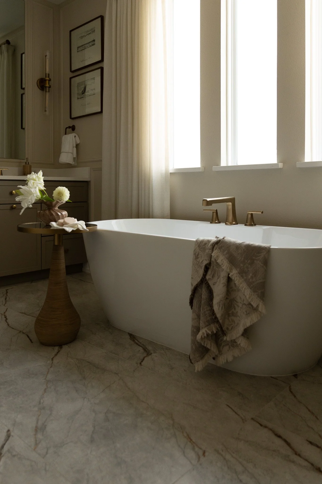 An elegant white bathtub with  soft light filtering in from the large window in the background. An accent table is in the front of the bathtub holding elegant bath accessories. 