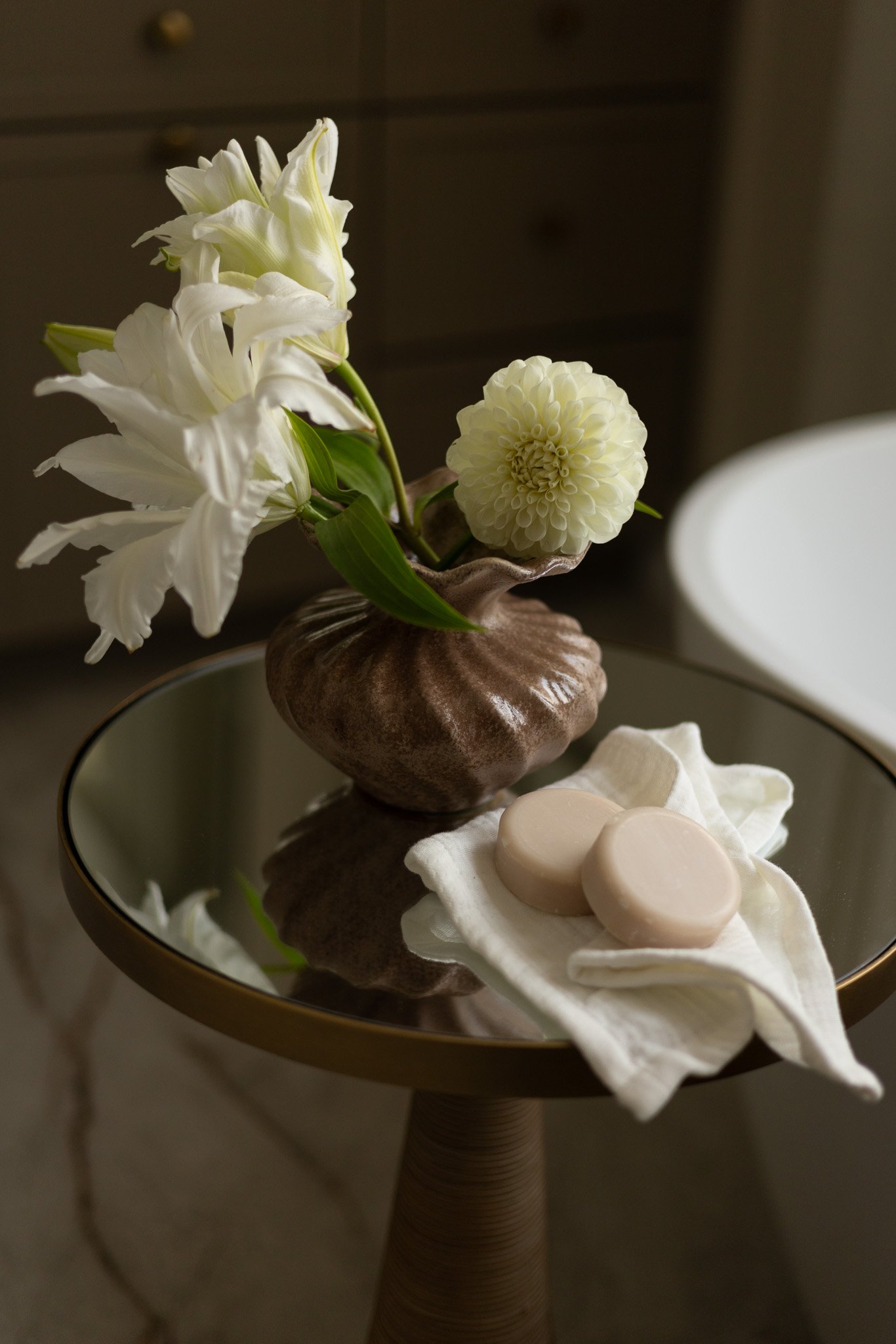 A close up shot of a vase with white flowers on a small side table next to the bathtub. A cloth with handmade soaps is placed next to the vase. 