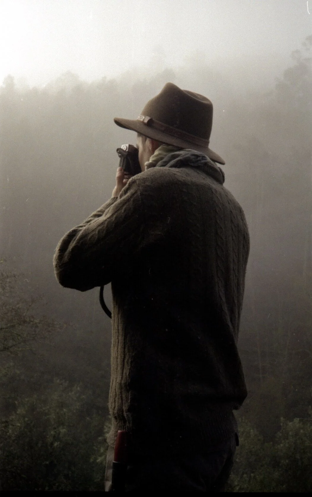 Pessoa com chapéu de bico largo, vestindo casaco de tricô, segurando uma câmera e apontando para frente, ao fundo uma paisagem escura com nuvens.