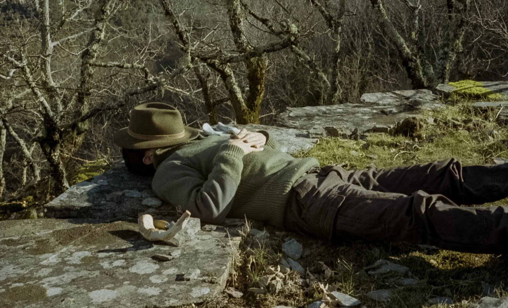 Pessoa deitada em uma rocha na natureza, usando chapéu e roupas de aventura, com árvores ao fundo.