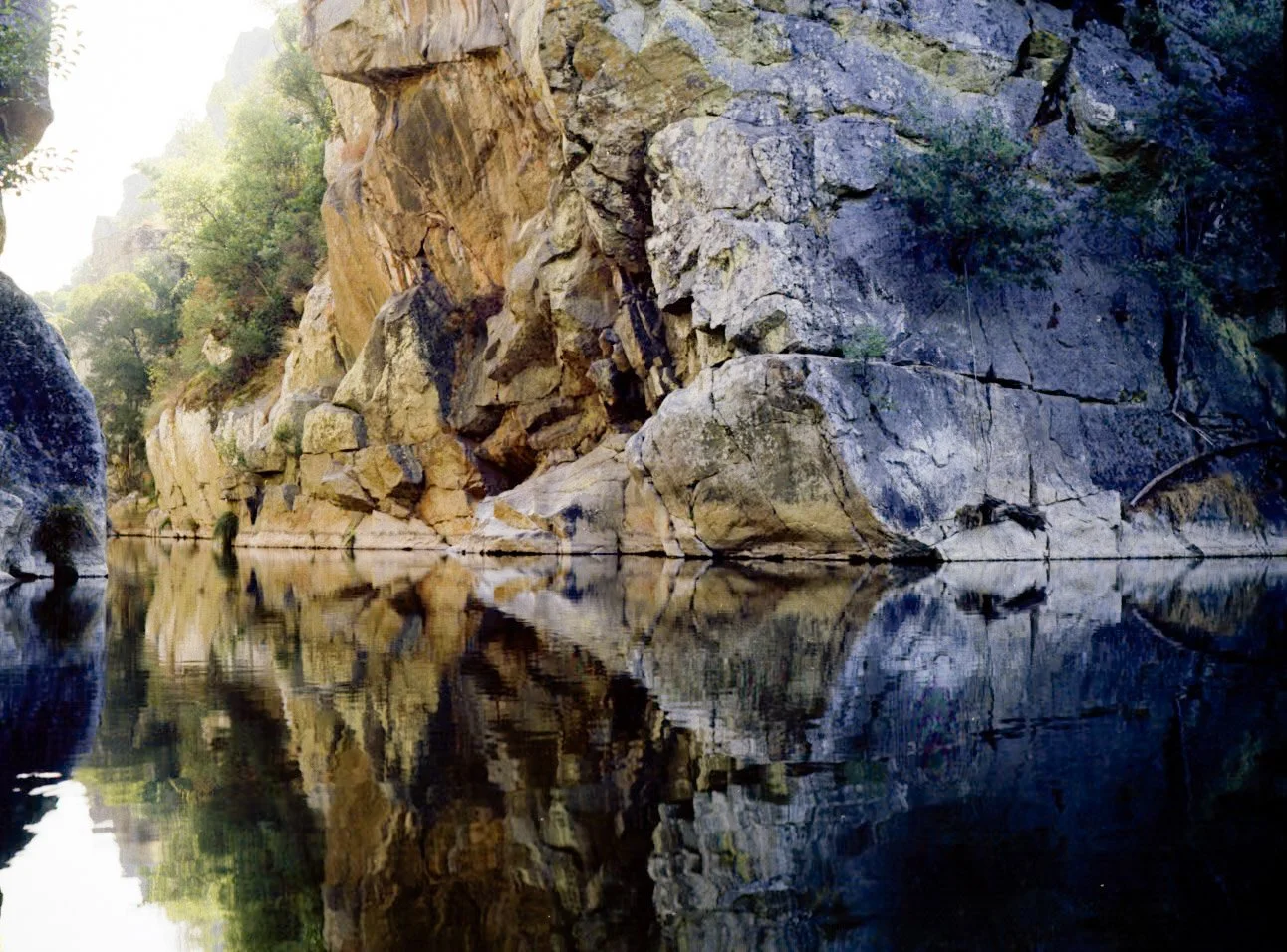 Cânion com grandes rochas de variadas cores refletidas em uma água tranquila, rodeado por vegetação.