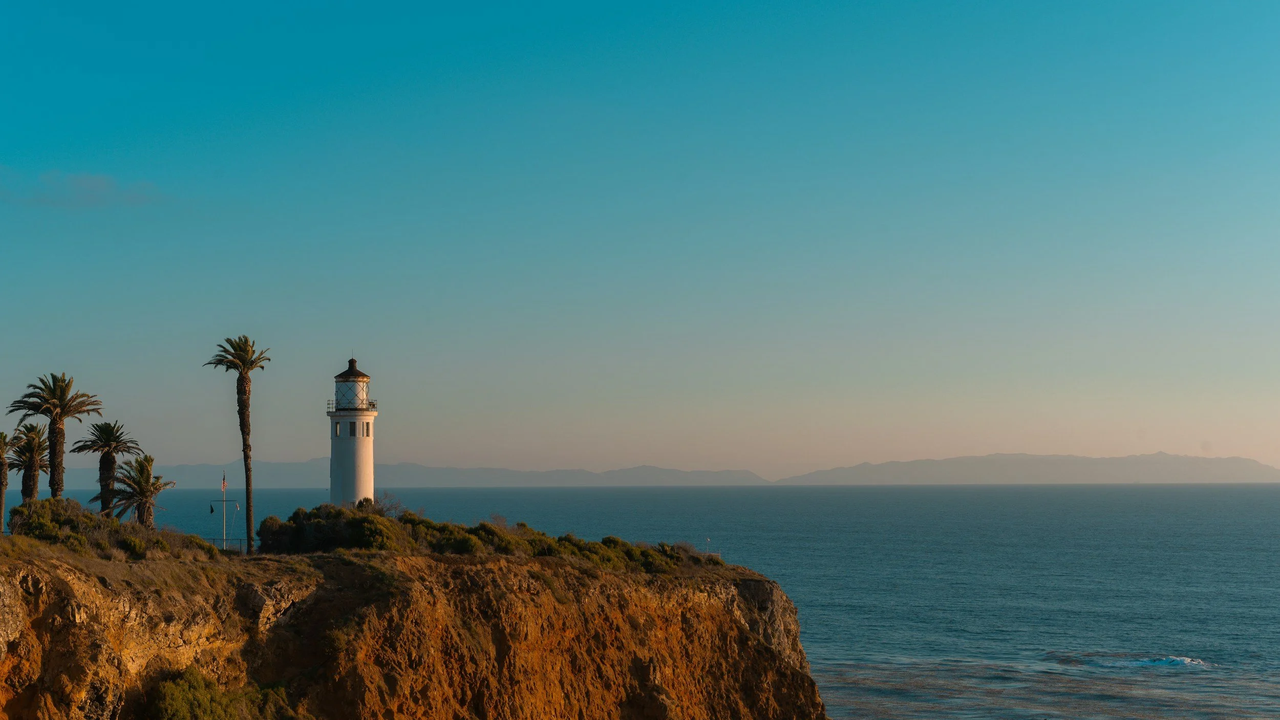 The Point Vicente lighthouse in Rancho Palos Verdes on the rocky cliff with palm trees, overlooking the ocean during sunset or sunrise, with distant mountains on the horizon.