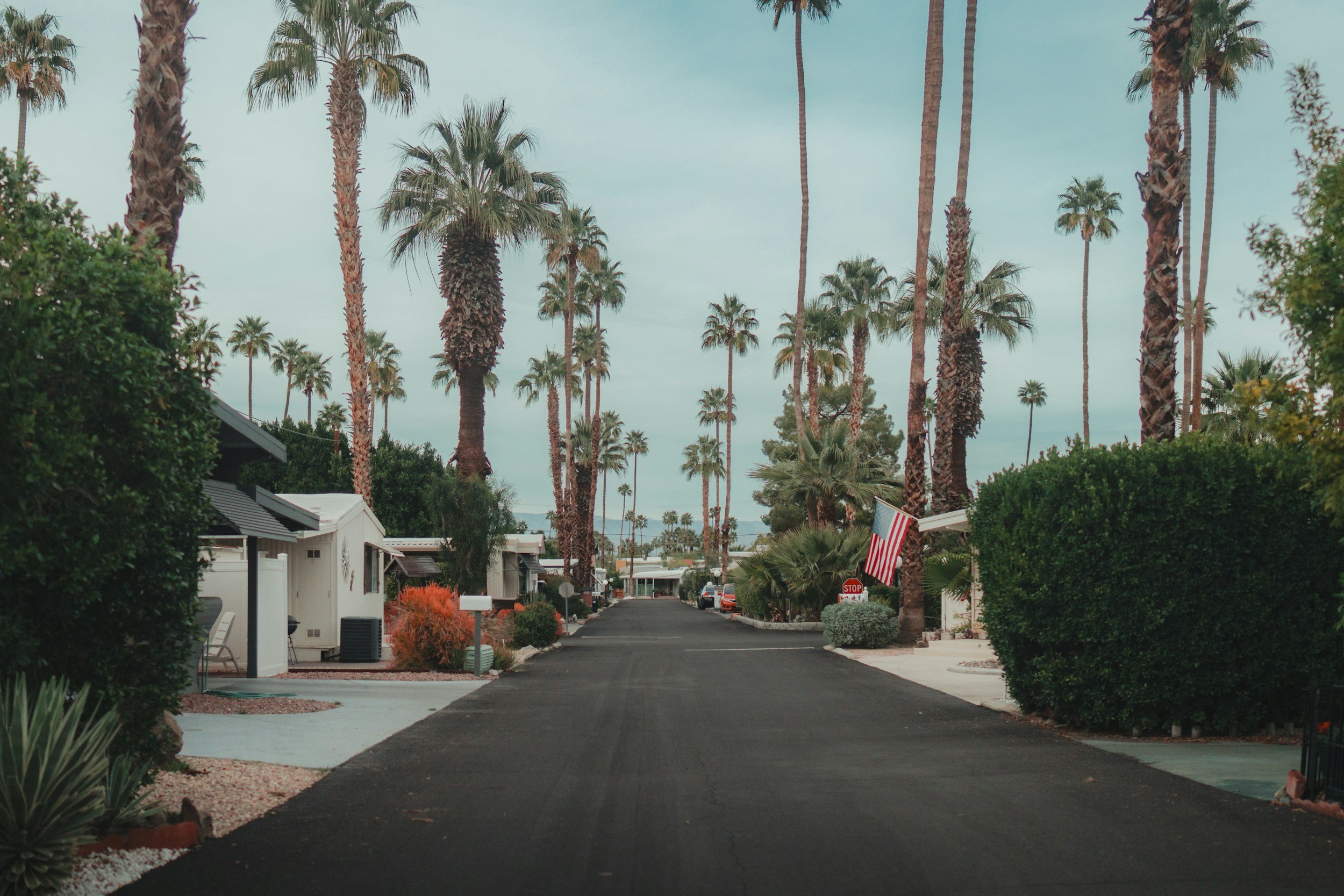 A Los Angeles house with a red-tiled roof and white stucco walls, surrounded by palm trees and lush greenery, under a partly cloudy blue sky.
