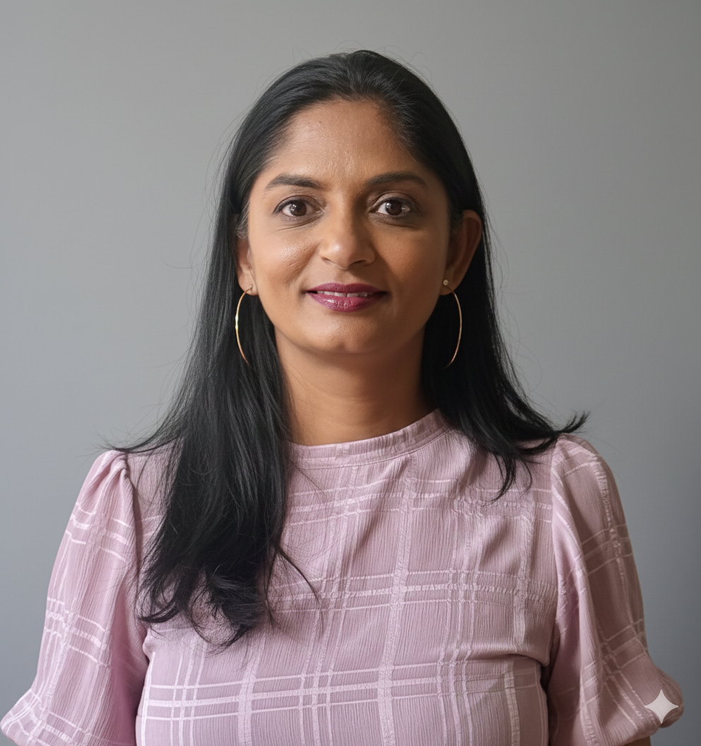 Sujana Patel wearing a pink blouse and hoop earrings, standing against a gray background.