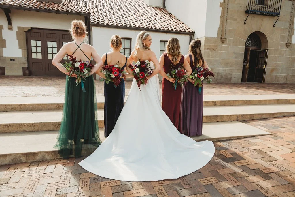 Bride in a white gown standing with four bridesmaids in colorful dresses, all holding bouquets of flowers, on steps outside a building with brick and stucco walls.
