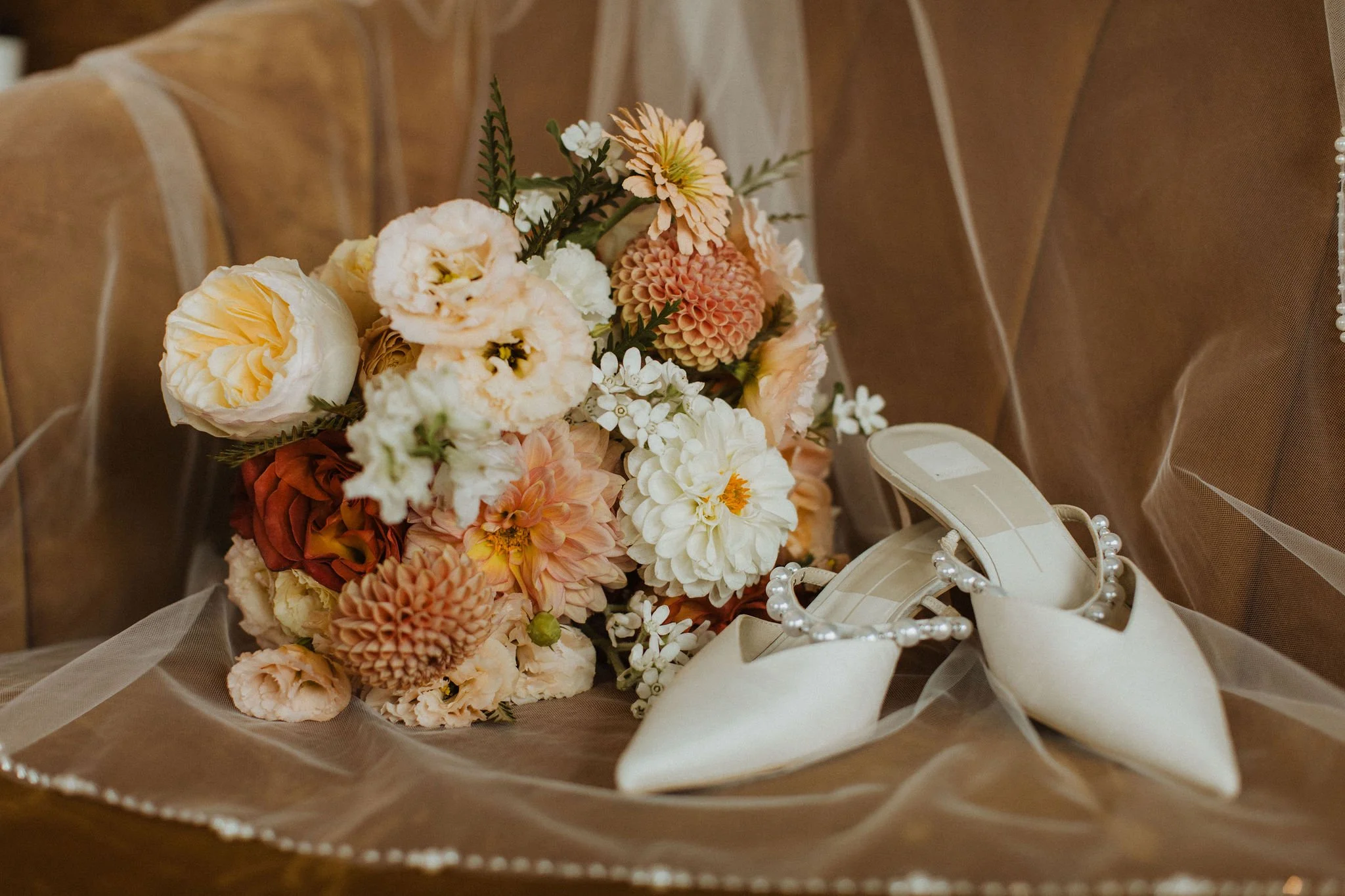 A bouquet of mixed flowers including white, peach, and pink blooms, placed on a table near a beige chair with a pearl necklace and a beige high heel shoe with a pearl detail.
