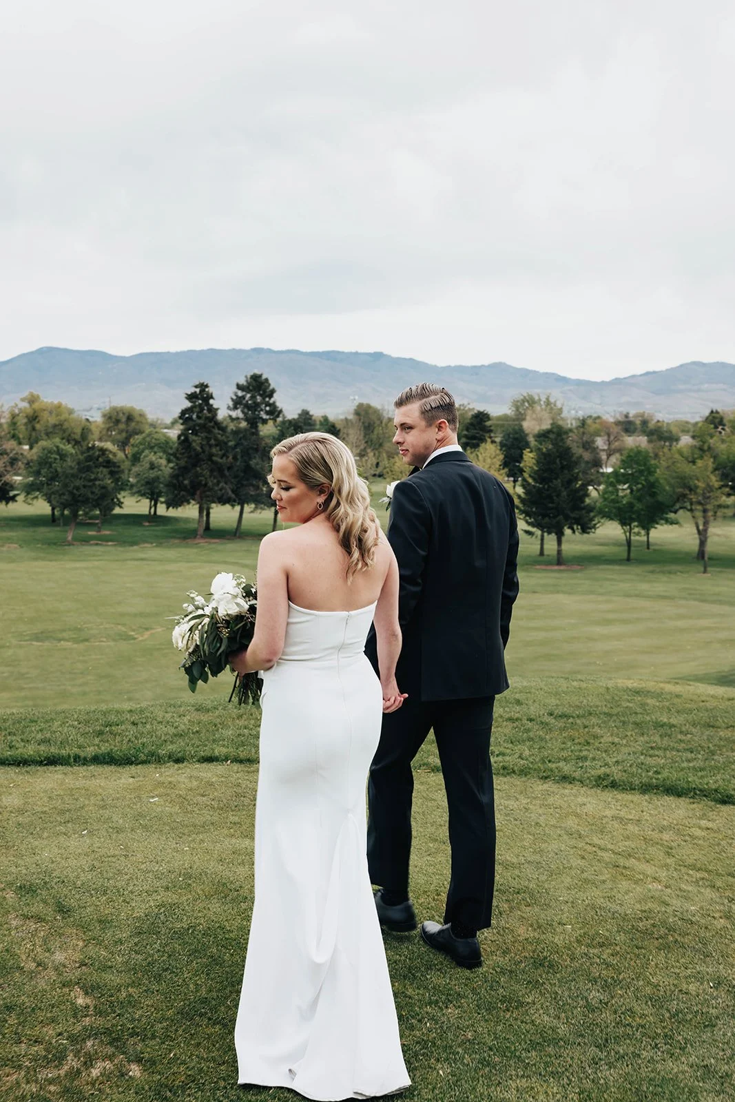 A bride in a strapless white wedding gown holding a bouquet, and a groom in a black suit holding hands, standing outdoors on a green field with trees and mountains in the background on a cloudy day.