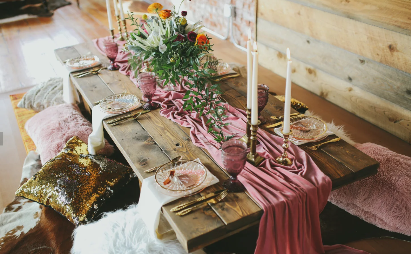 A rustic wooden dining table decorated for a celebration with a pink table runner, tall candles, pink and purple glassware, floral centerpiece, and plush pink and sequin pillows around the seating area.