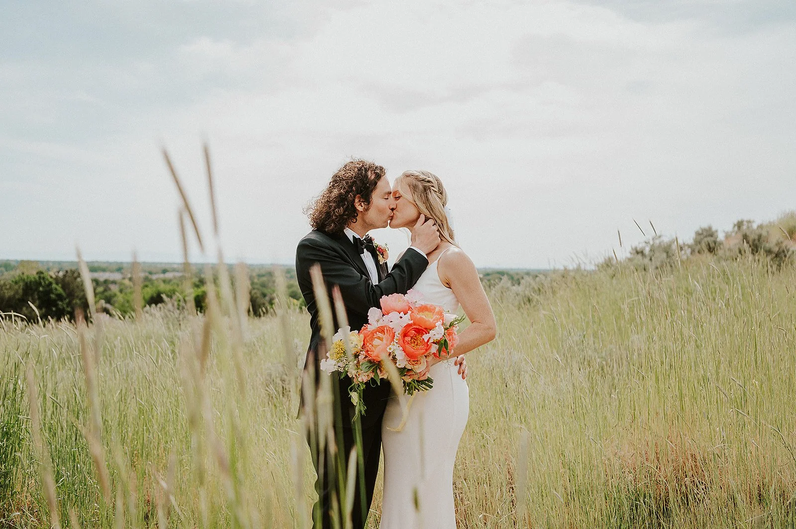 A bride and groom kiss in a grassy field, with the groom in a tuxedo holding a bouquet, and the bride in a white wedding dress with a pink and orange floral bouquet.
