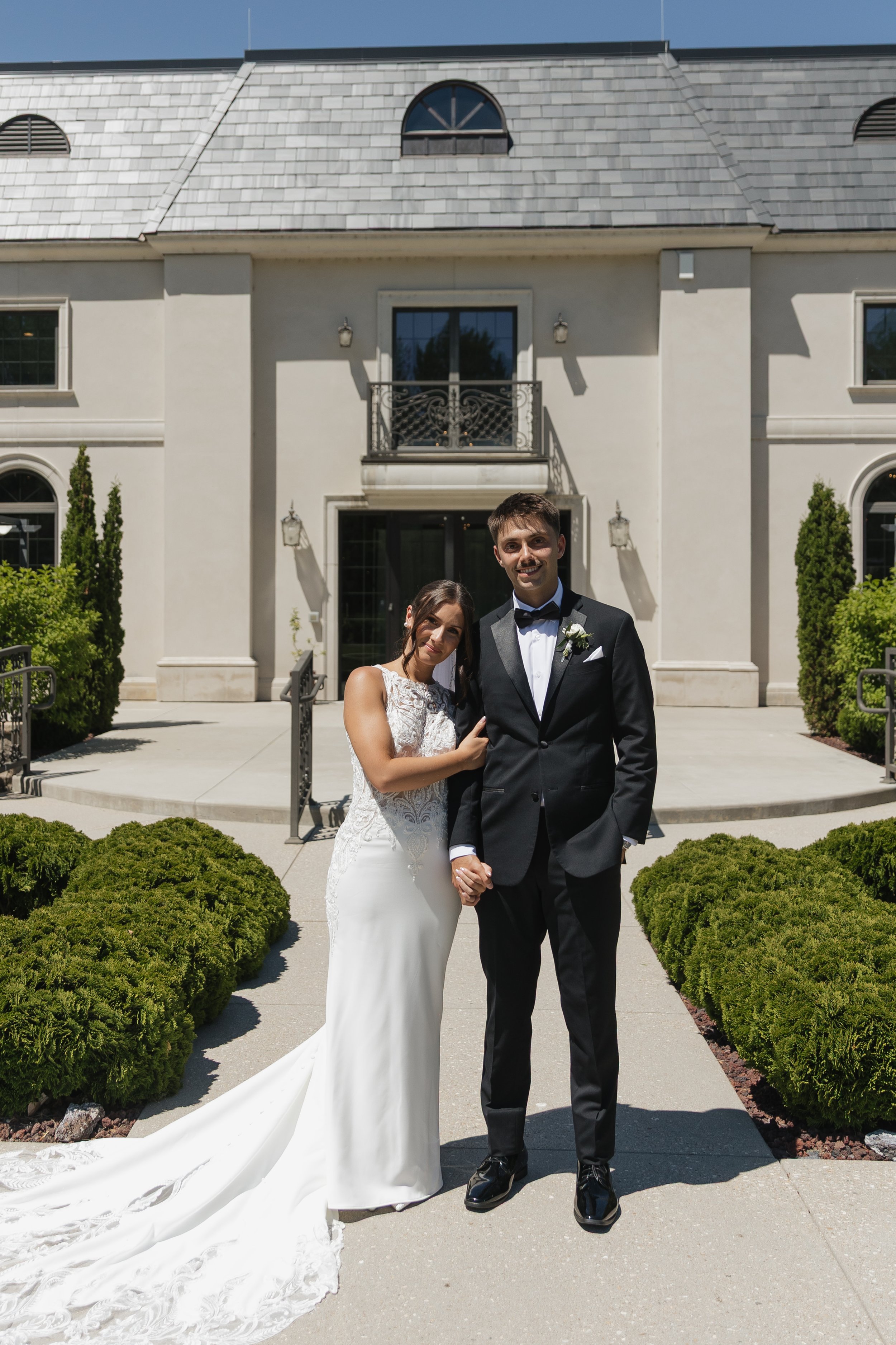 A bride and groom standing outdoors in front of a large, white, elegant building, holding hands and smiling. The bride is wearing a white wedding gown with lace details, and the groom is dressed in a black tuxedo with a bow tie and a white boutonniere. There is greenery and bushes surrounding them, with clear skies above.