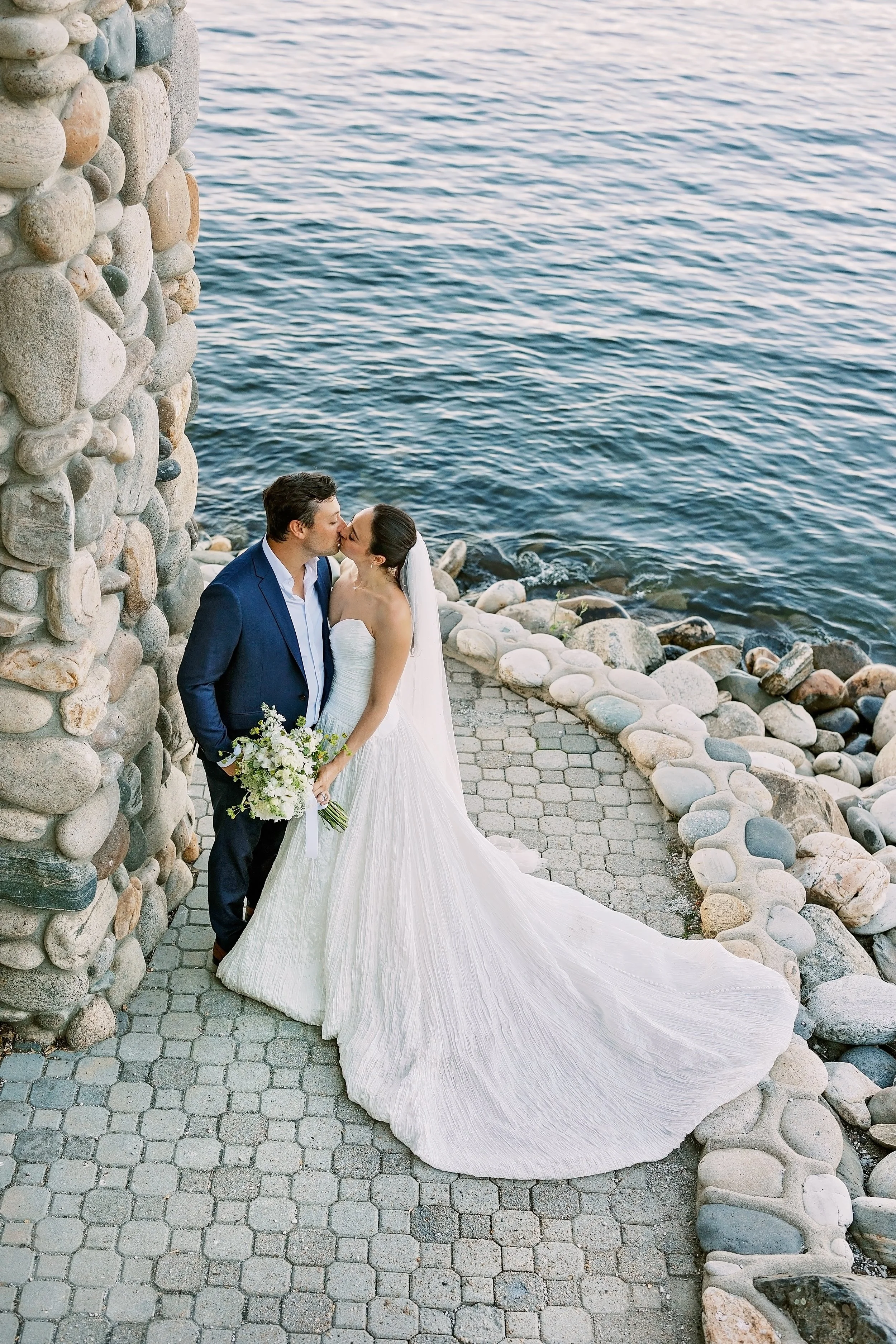 A bride and groom kissing by a rocky shoreline, with a stone wall on the left side and ocean water in the background.