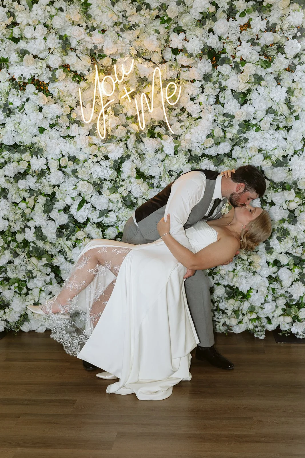 A bride and groom sharing a kiss in front of a wall covered with white flowers and a neon sign that reads 'you + me'.