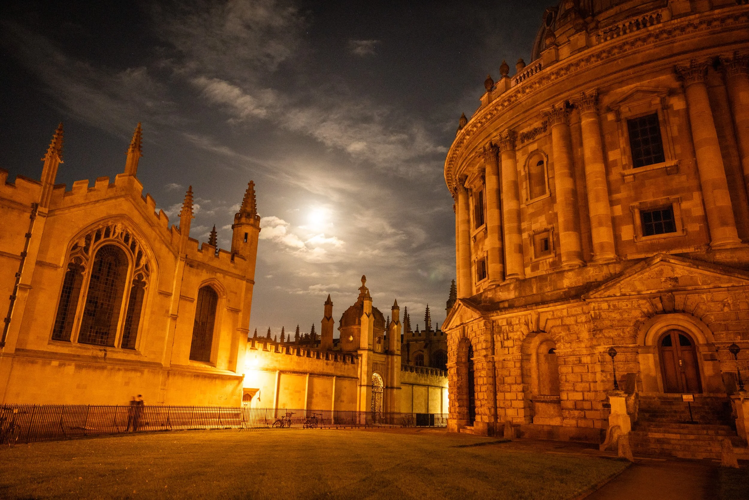 Nighttime view of historic Oxford University with the Radcliffe Camera in the foreground and All Souls College in the background, all illuminated by warm exterior lights, under a moonlit sky with scattered clouds.