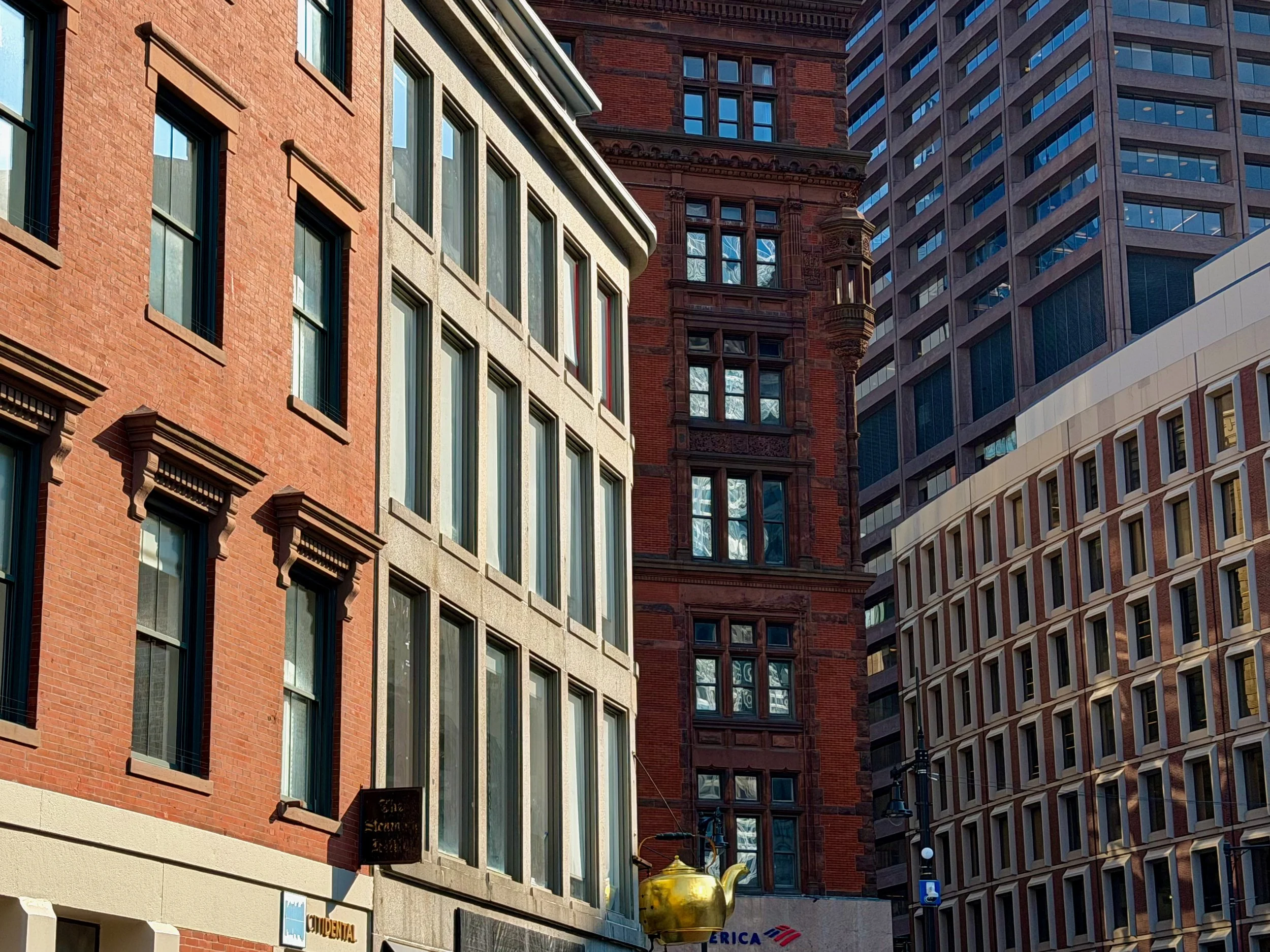 A Boston city street with tall buildings of different architectural styles, including a red brick building and modern glass skyscrapers. A yellow teapot decoration is visible outside a building at street level.