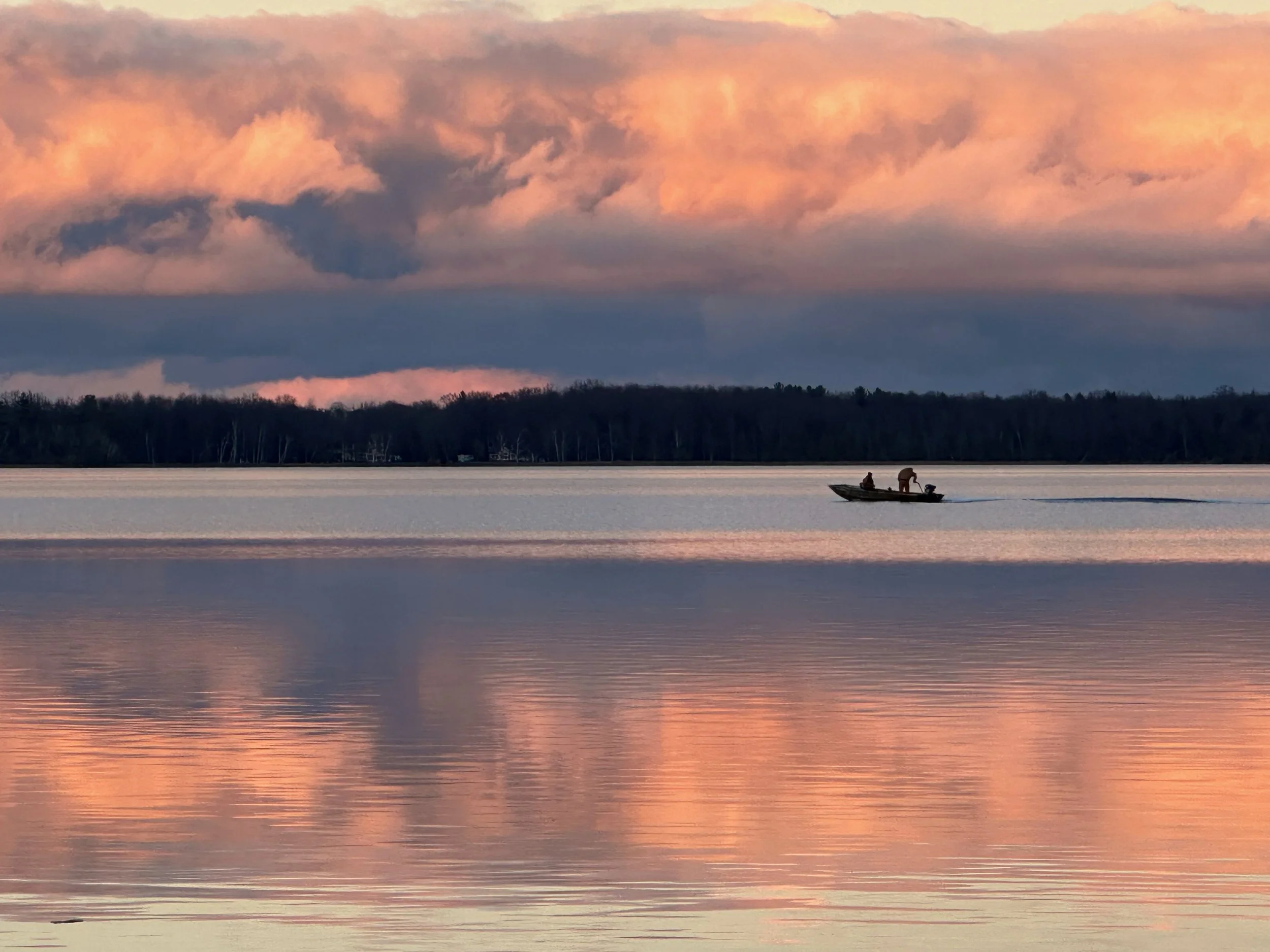 A boat with two people on a lake during sunset, with pink and orange clouds in the sky and trees along the shoreline.