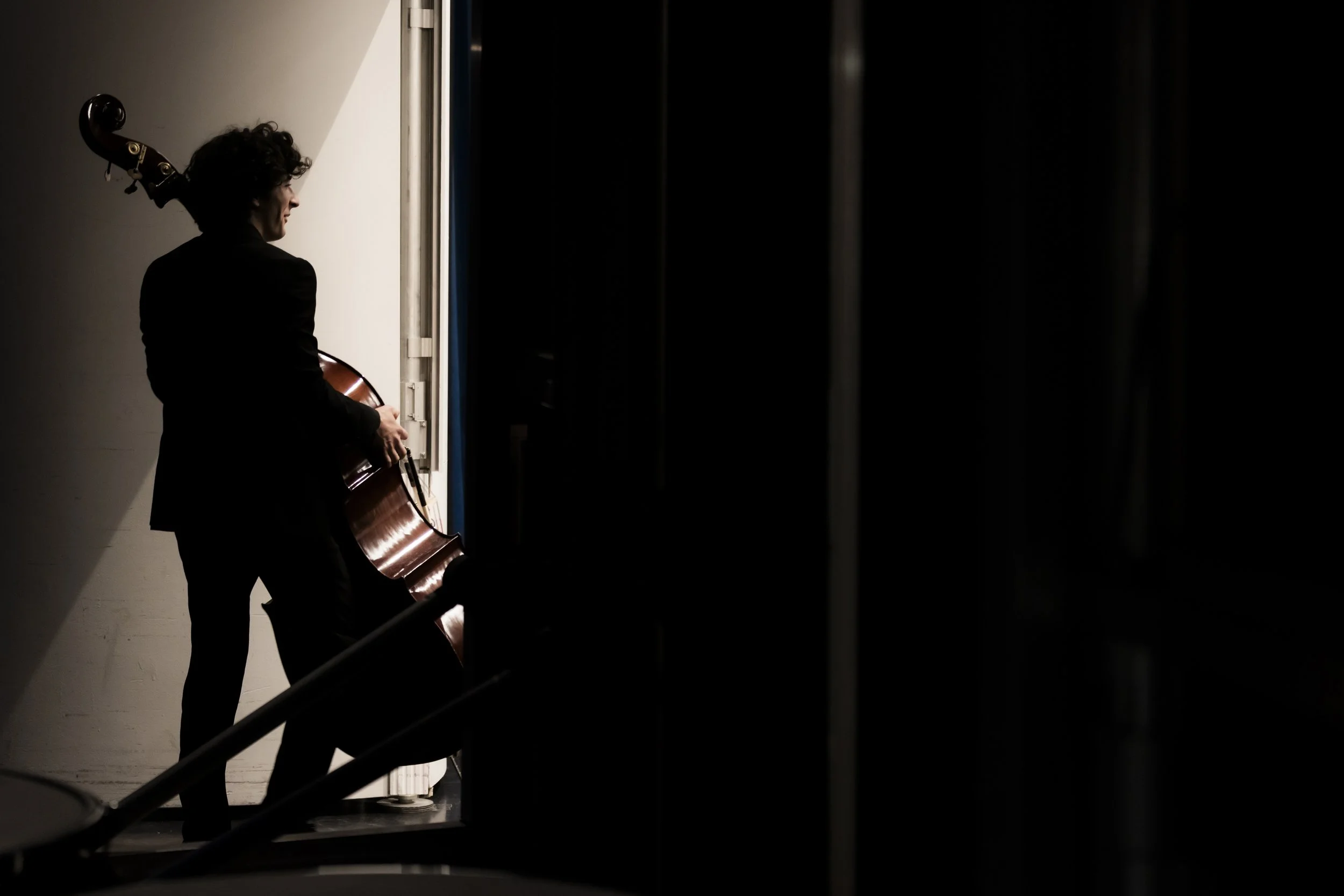 A person with curly hair playing a cello backstage, silhouetted against a starkly lit wall, with dim surroundings and a dark background.