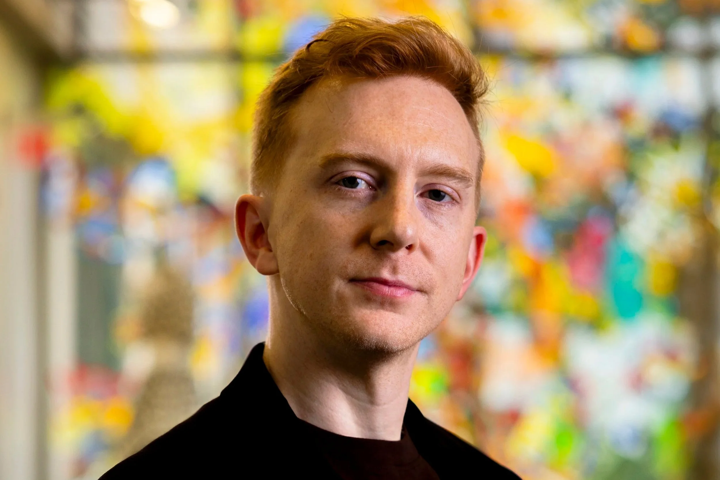 A young man with red hair and blue eyes standing indoors with colorful blurred decorations in the background.
