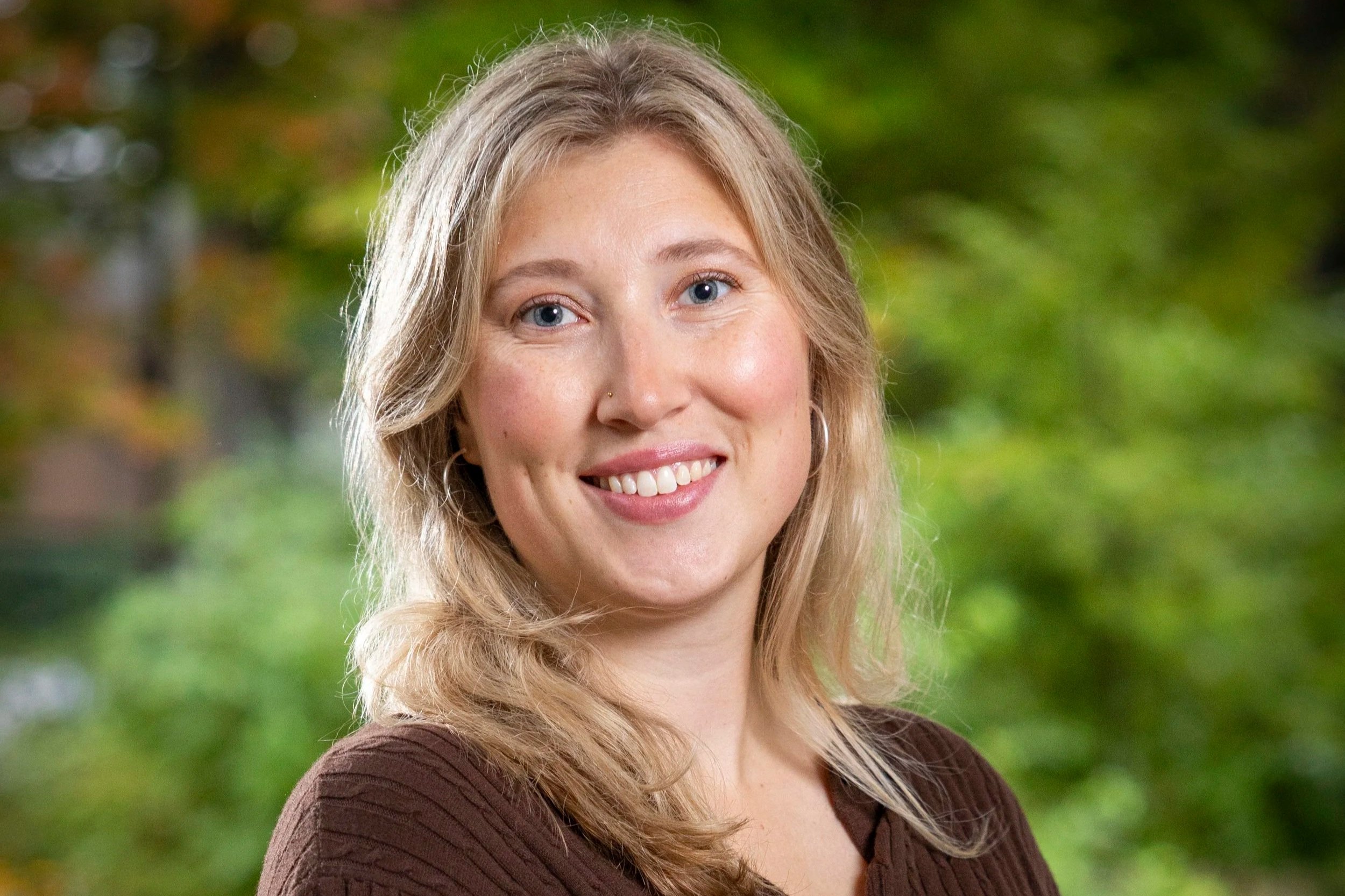Close-up of a woman with blonde hair, blue eyes, and light skin, smiling outdoors with a blurred green background.