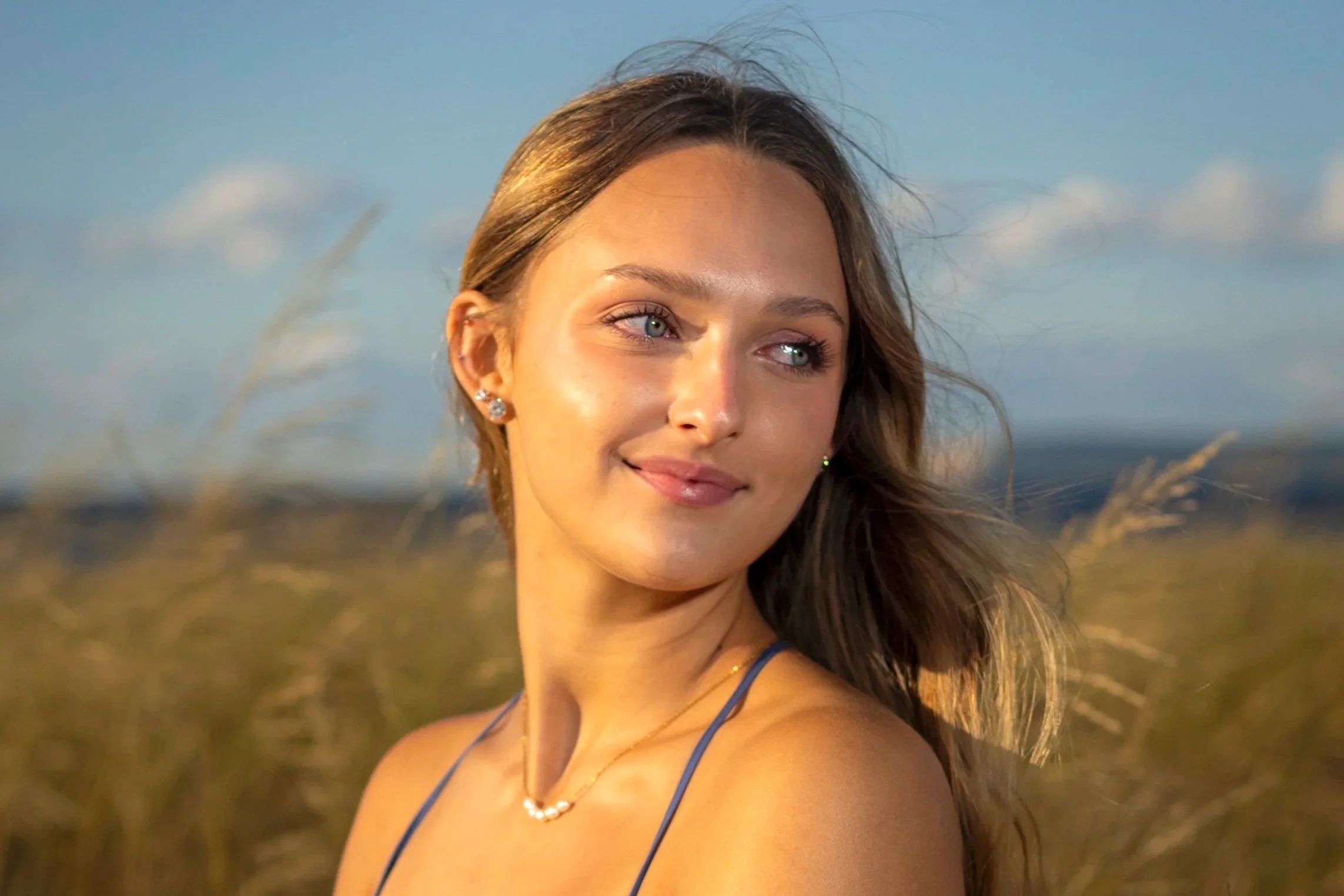Close-up of a woman with blue eyes, long brown hair, wearing earrings and a pearl necklace, outdoors in sunlight, with a blurred natural background