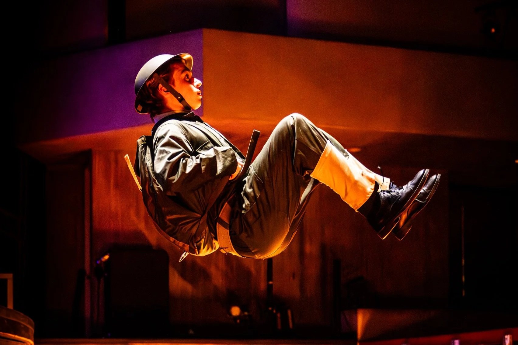 An actor in the musical "Tommy" wearing a soldier's uniform jumps from the stage and appears floating or suspended in a low-light indoor environment with warm, reddish lighting.