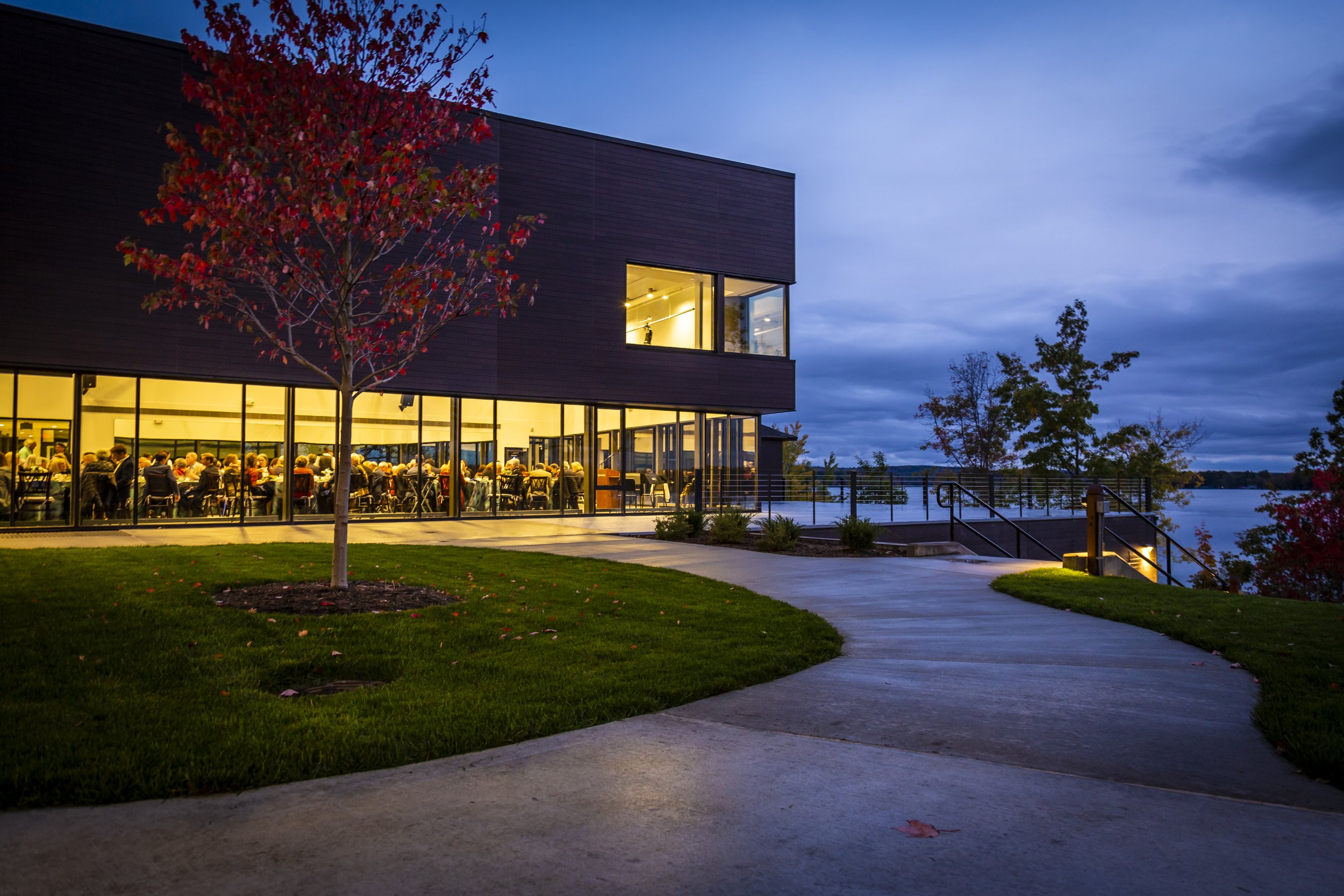 Modern building with large glass windows illuminated from inside, surrounded by trees with autumn leaves, by a waterfront with a walking path in the evening.