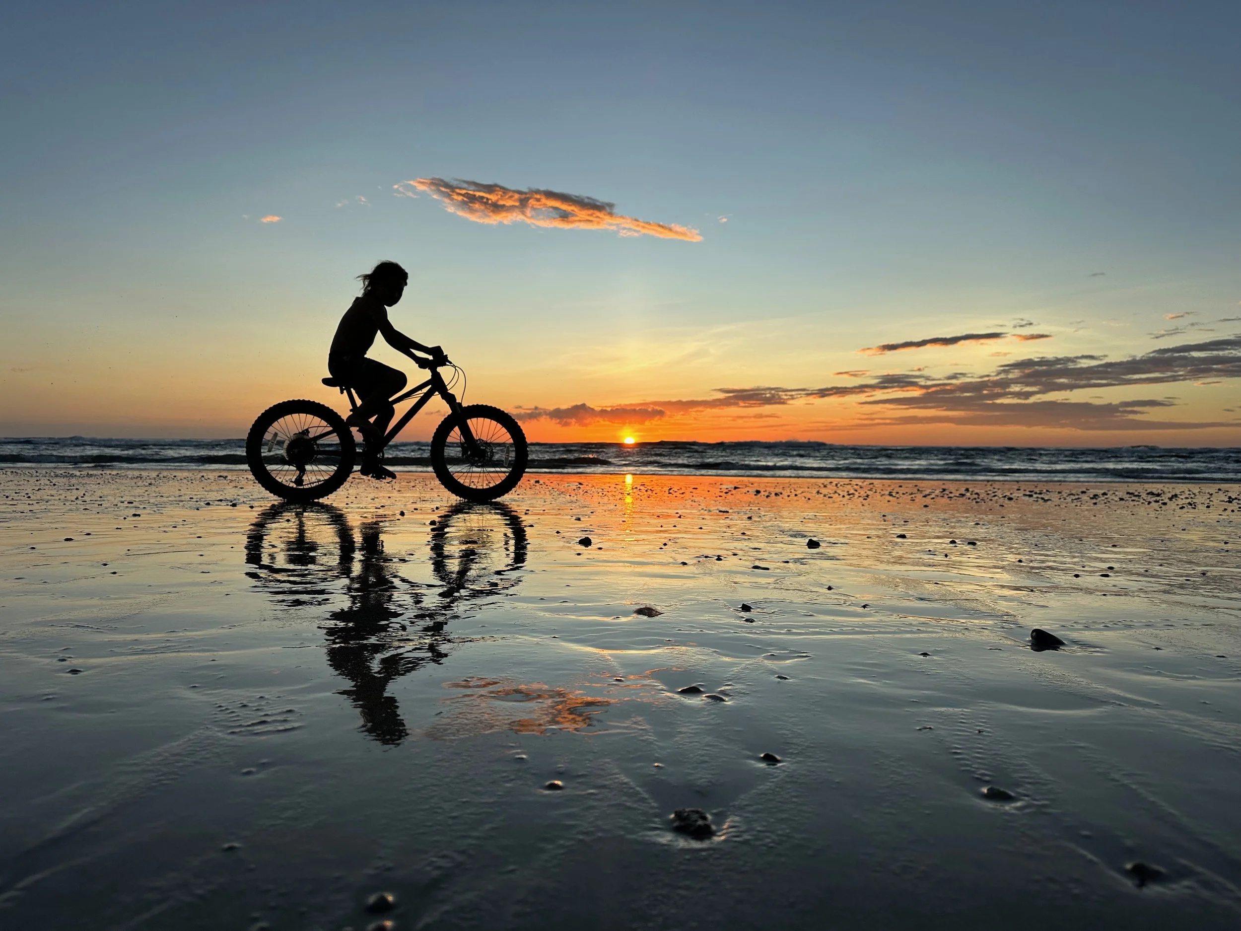 Silhouette of a person riding a bicycle on a beach during sunset, with the sun near the horizon and some clouds in the sky.