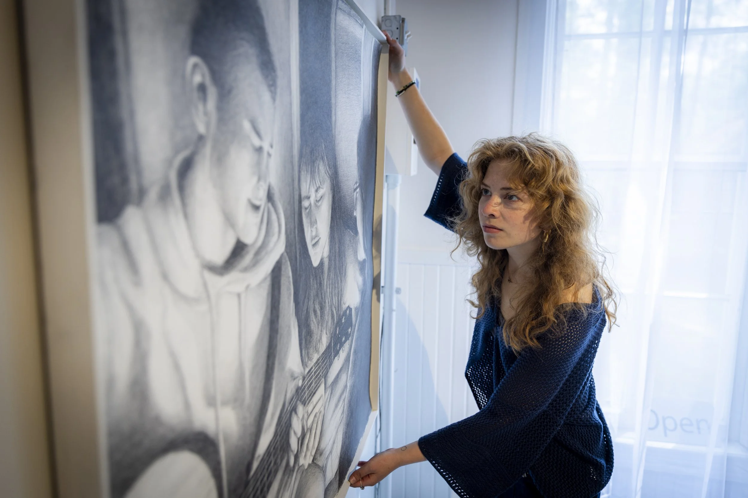 A woman with curly hair and a blue sweater is hanging a framed black and white drawing on a wall in a well-lit room with white curtains.