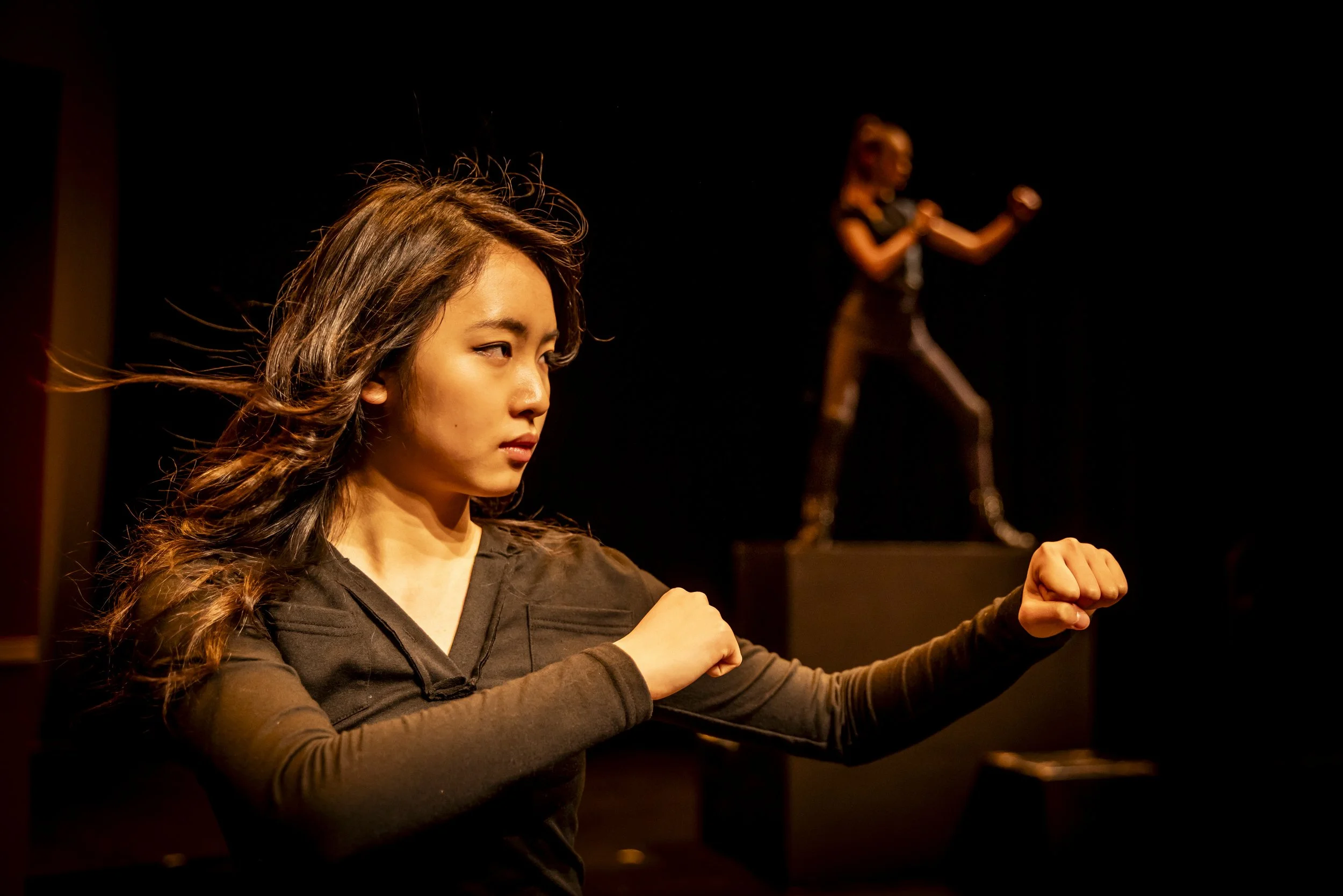 A woman with long, wavy hair and a serious expression, wearing a dark long-sleeve shirt, raising her fists in a fighting stance on stage with a dark background. Another woman is blurred in the background, also on stage.