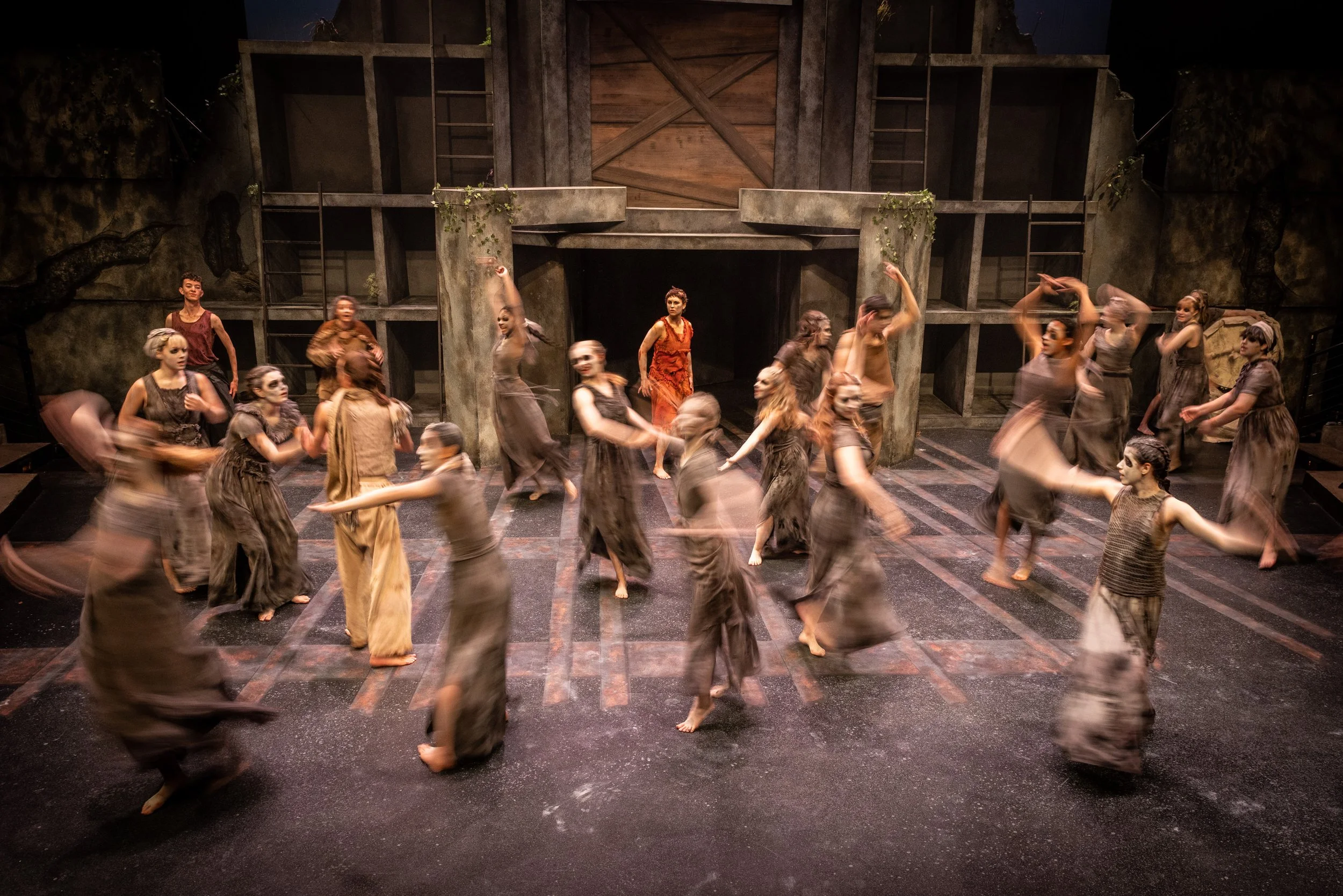 A theatrical stage with a group of performers dressed in costumes moving in a dance or choreographed scene in front of a set designed to look like a rustic or ancient structure.