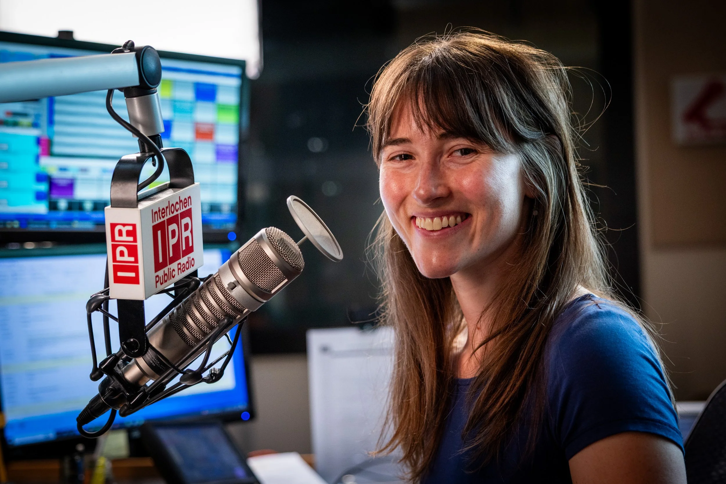 Smiling woman in a radio studio, speaking into a microphone with a "Public Radio" label, in front of a computer monitor with colorful waveforms.