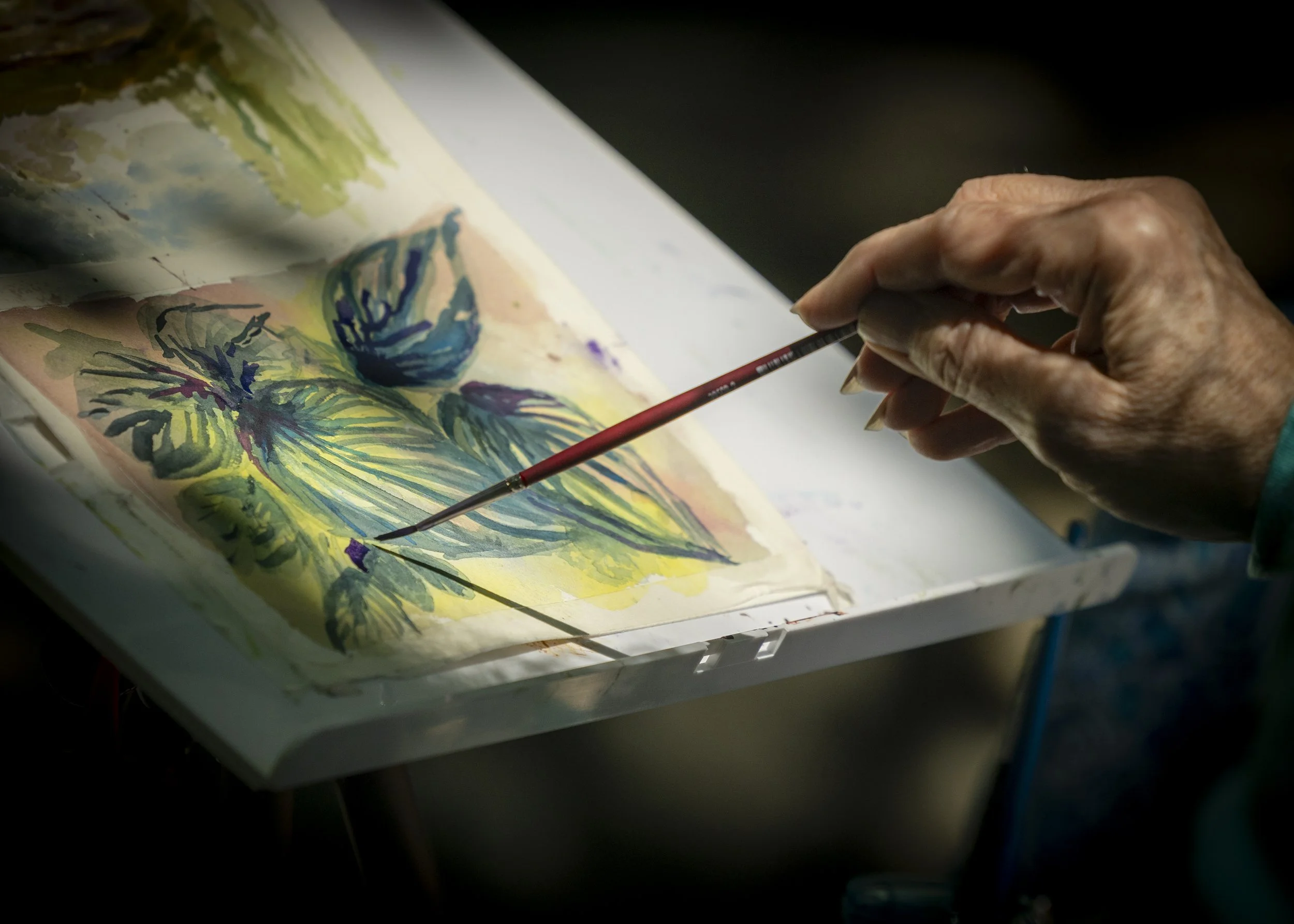 Close-up of a person's hand holding a thin paintbrush, painting a watercolor landscape with trees, a house, and a pool.