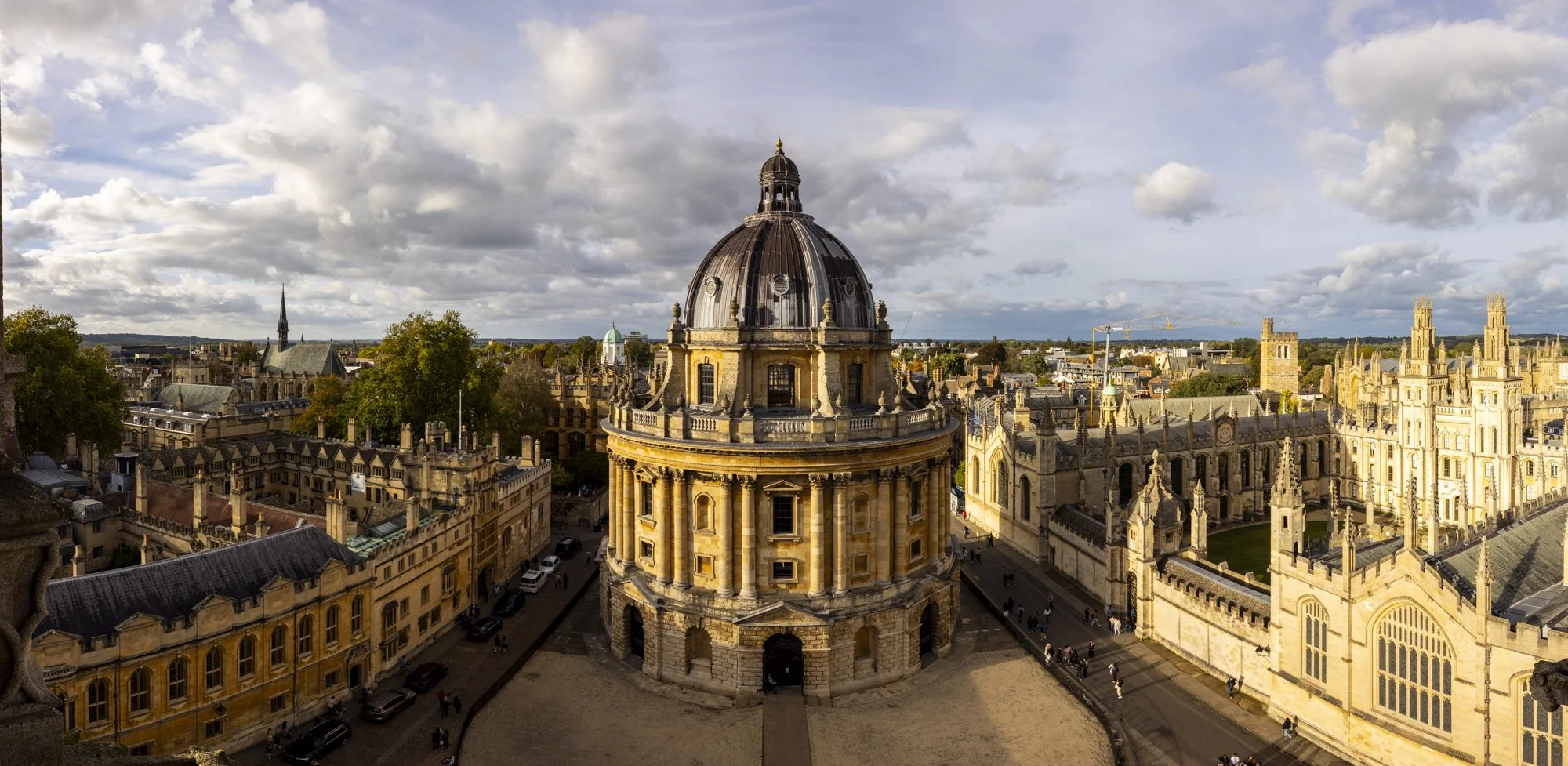 View of historic Oxford skyline showing Radcliffe Camera, surrounding university buildings, and a partly cloudy sky.