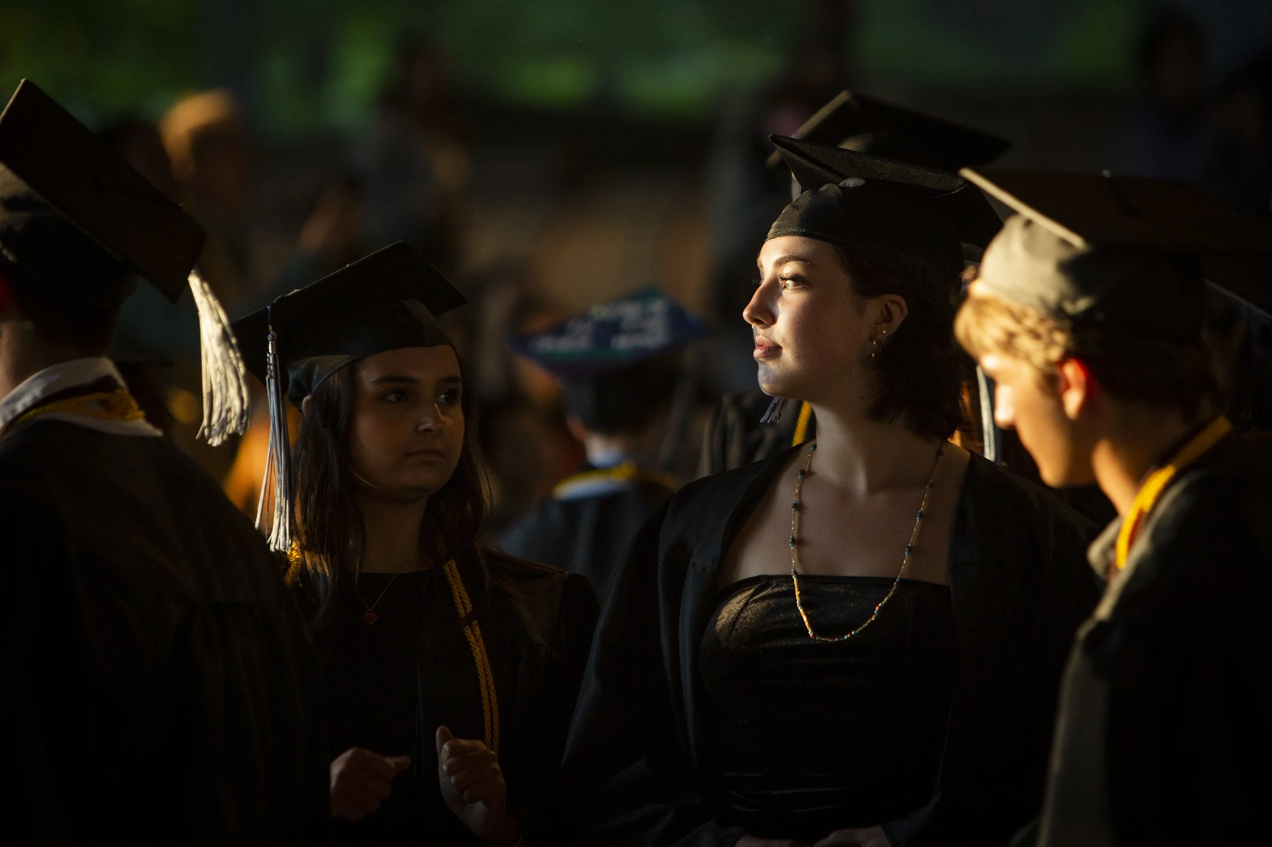 Group of diverse young adults wearing graduation caps and gowns at a graduation ceremony, as sunlight catches a young woman's face. 