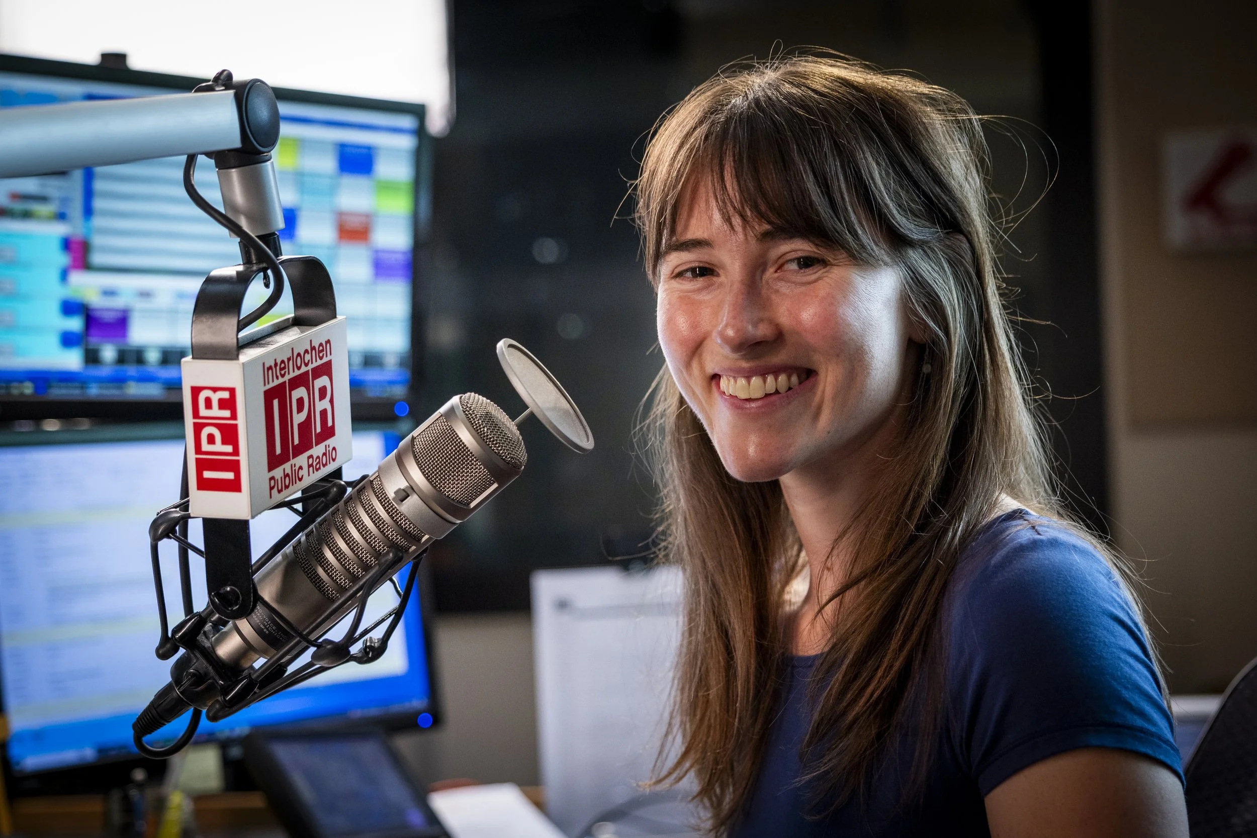 A woman with long brown hair and a blue shirt sitting in a radio studio with a microphone, computer screens, and radio station branding.