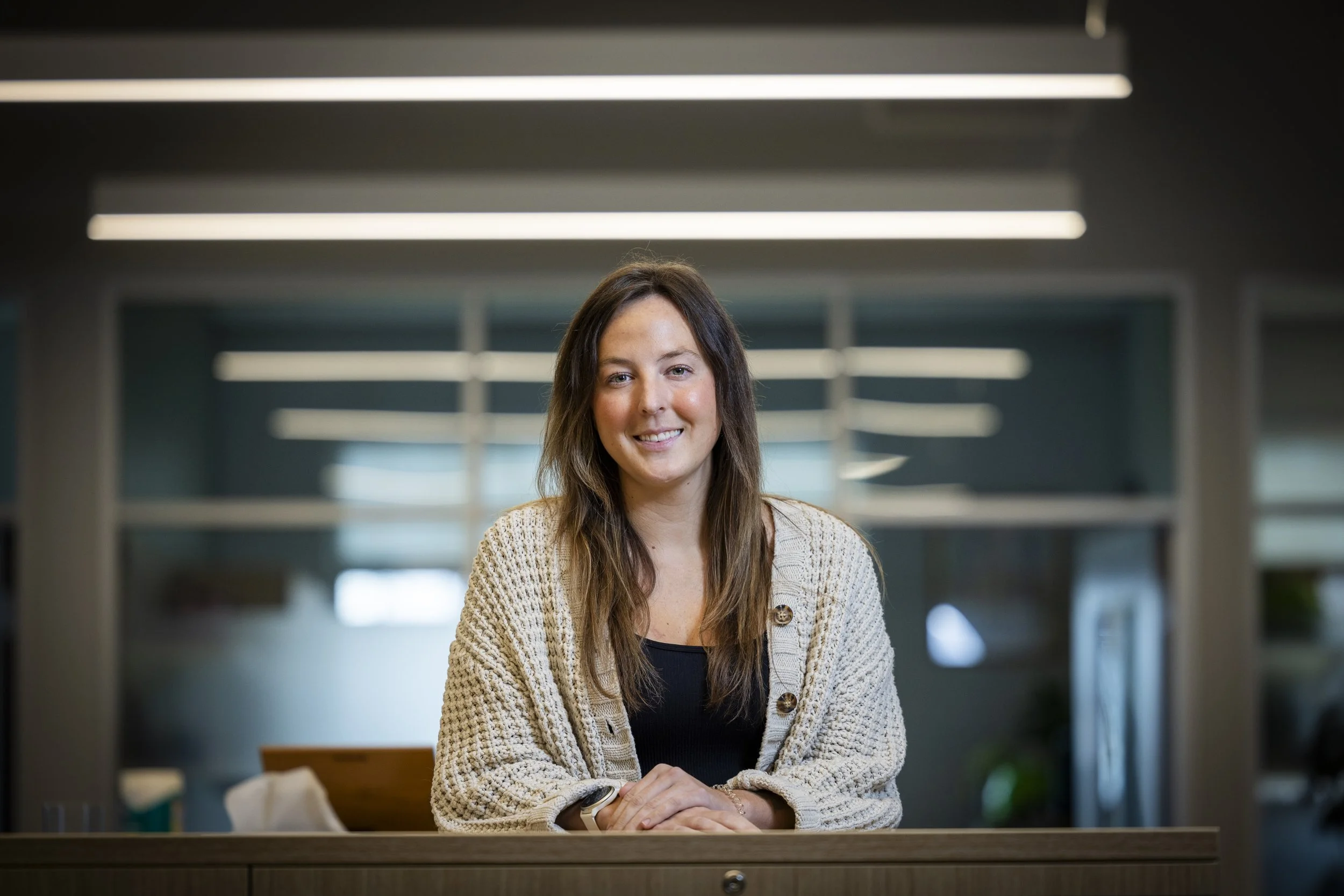 A young woman with long brown hair smiling, sitting at a desk in a modern office environment with fluorescent lighting.
