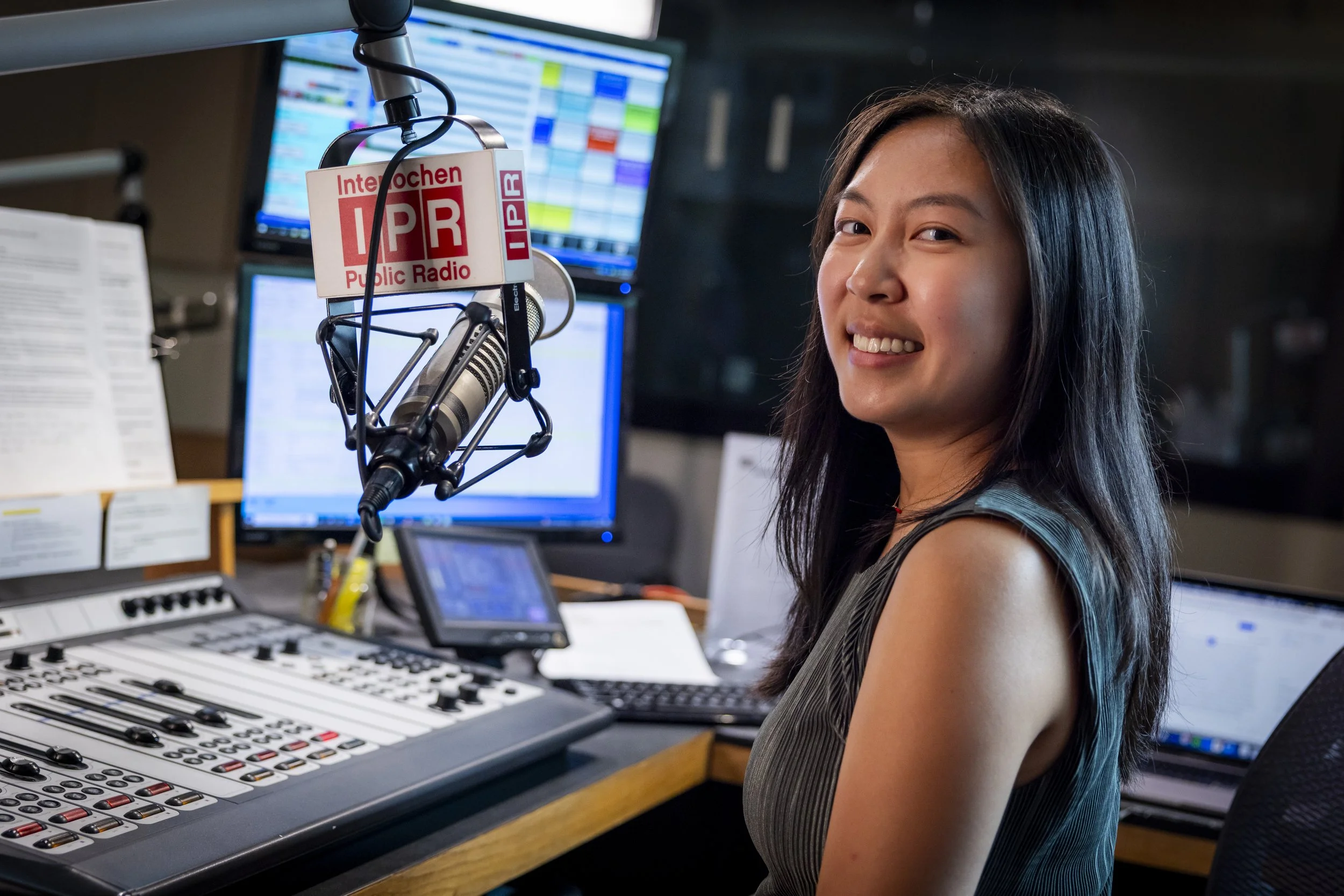 A woman smiling in a radio studio with a microphone that has a logo reading 'Inochen NPR Public Radio', and radio and computer equipment in the background.