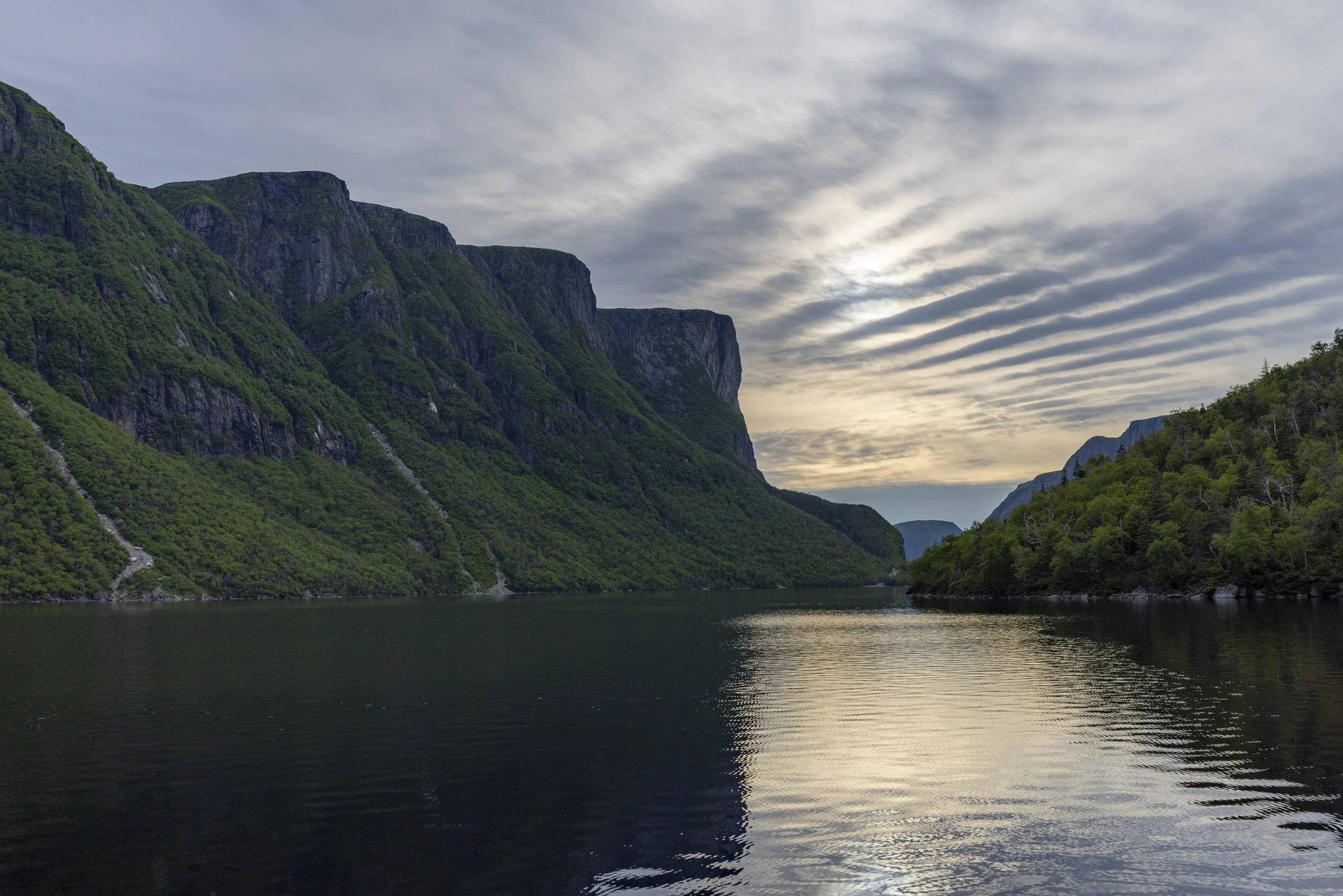 Scenic view of green mountains and cliffs with a body of water in the foreground, under a partly cloudy sky.