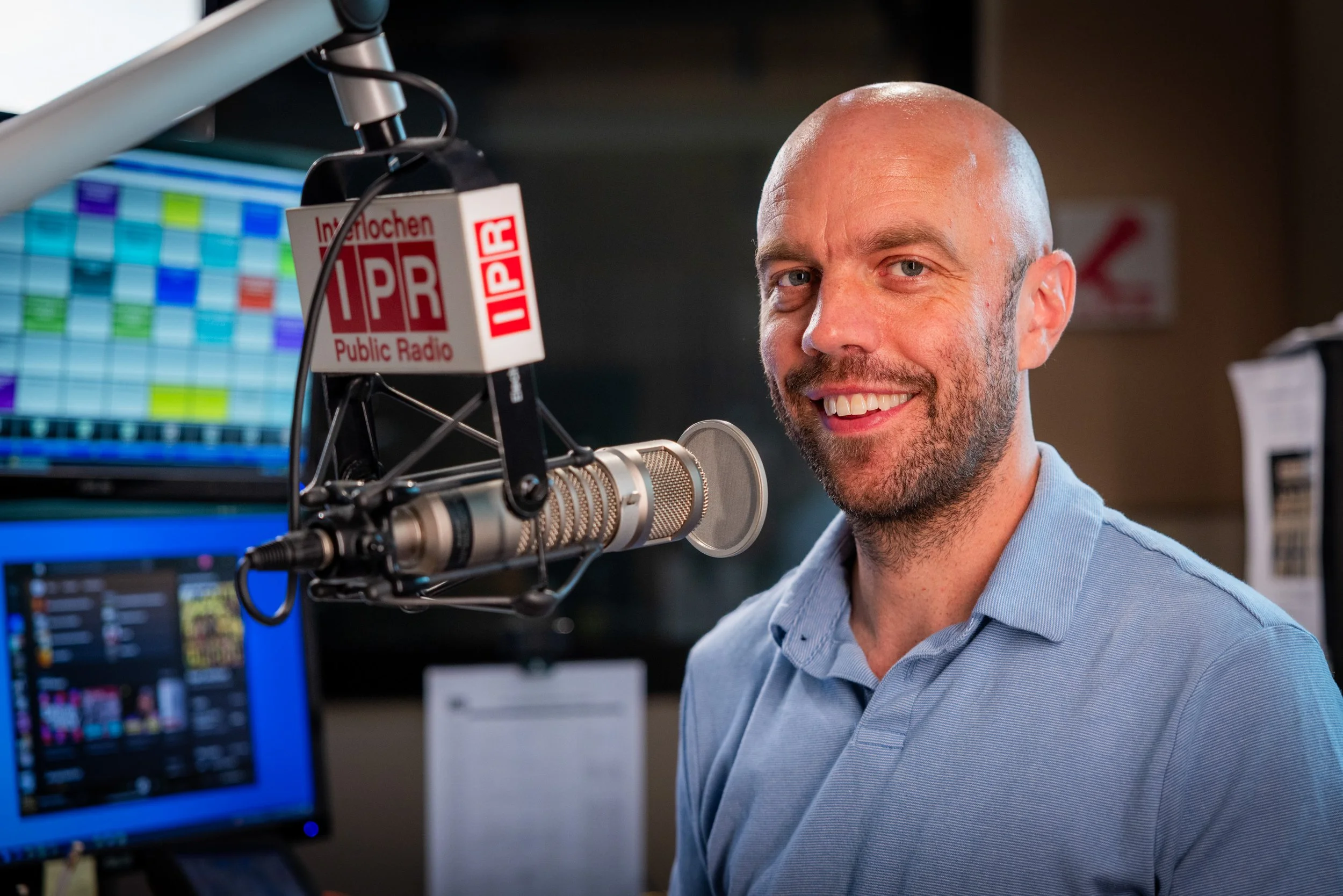 A man with a shaved head and beard smiling into a professional microphone in a radio station studio with colorful computer screens in the background.
