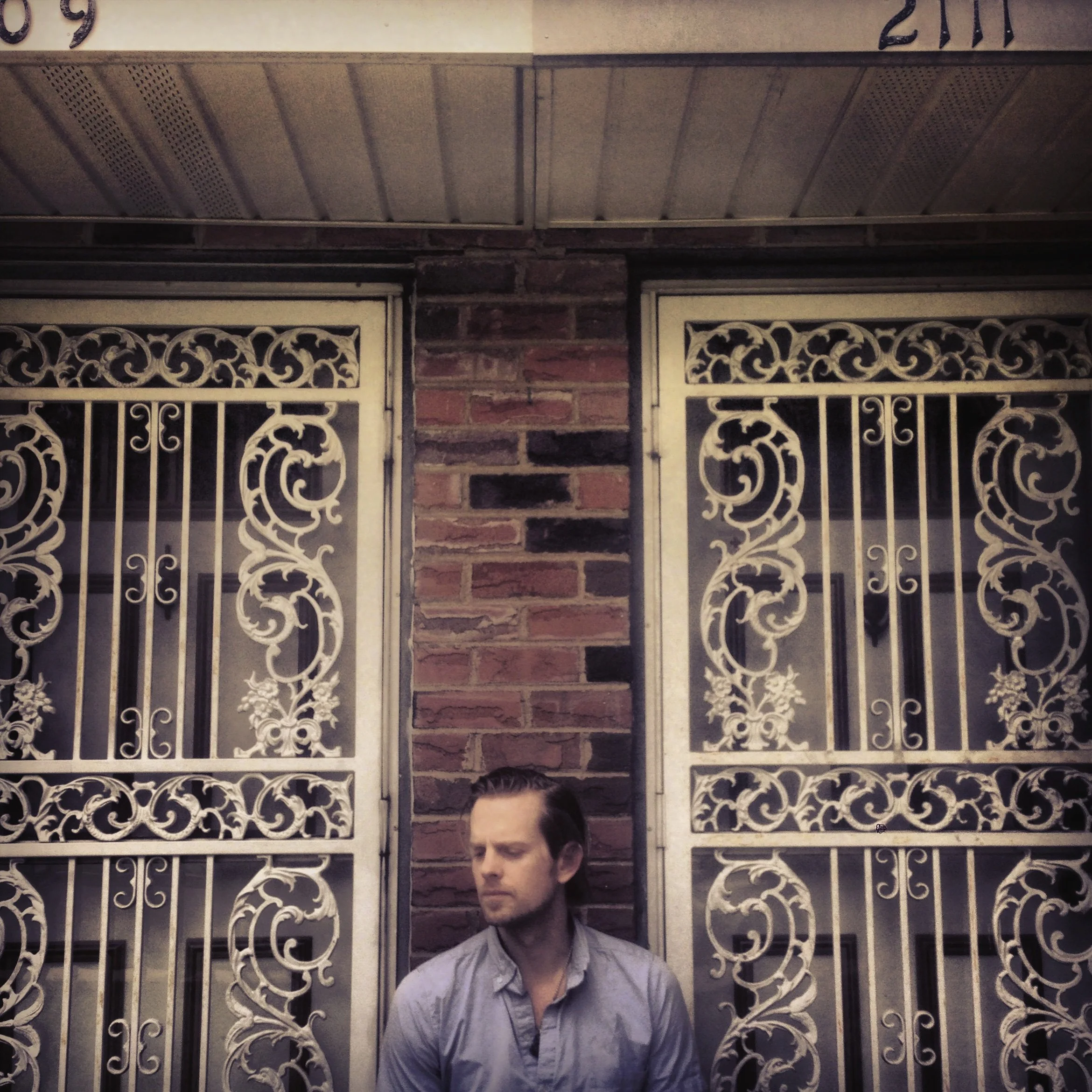 A man with short slicked-back hair and a beard stands in front of ornate white metal gates with intricate scrollwork design, next to a brick wall