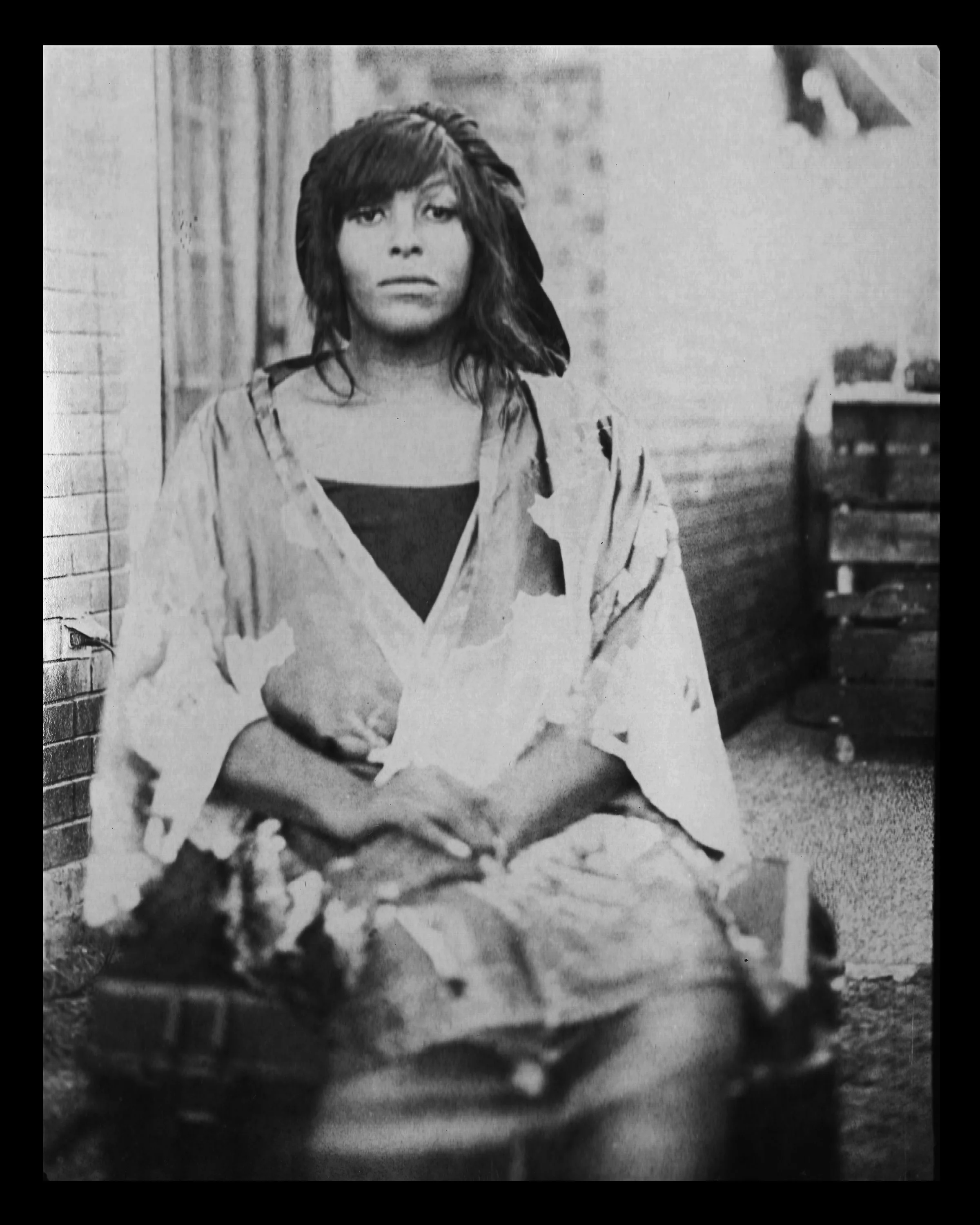 Black and white photo of a woman with disheveled hair sitting on the ground, looking directly at the camera with a serious expression. She wears a torn shirt and is surrounded by urban debris.