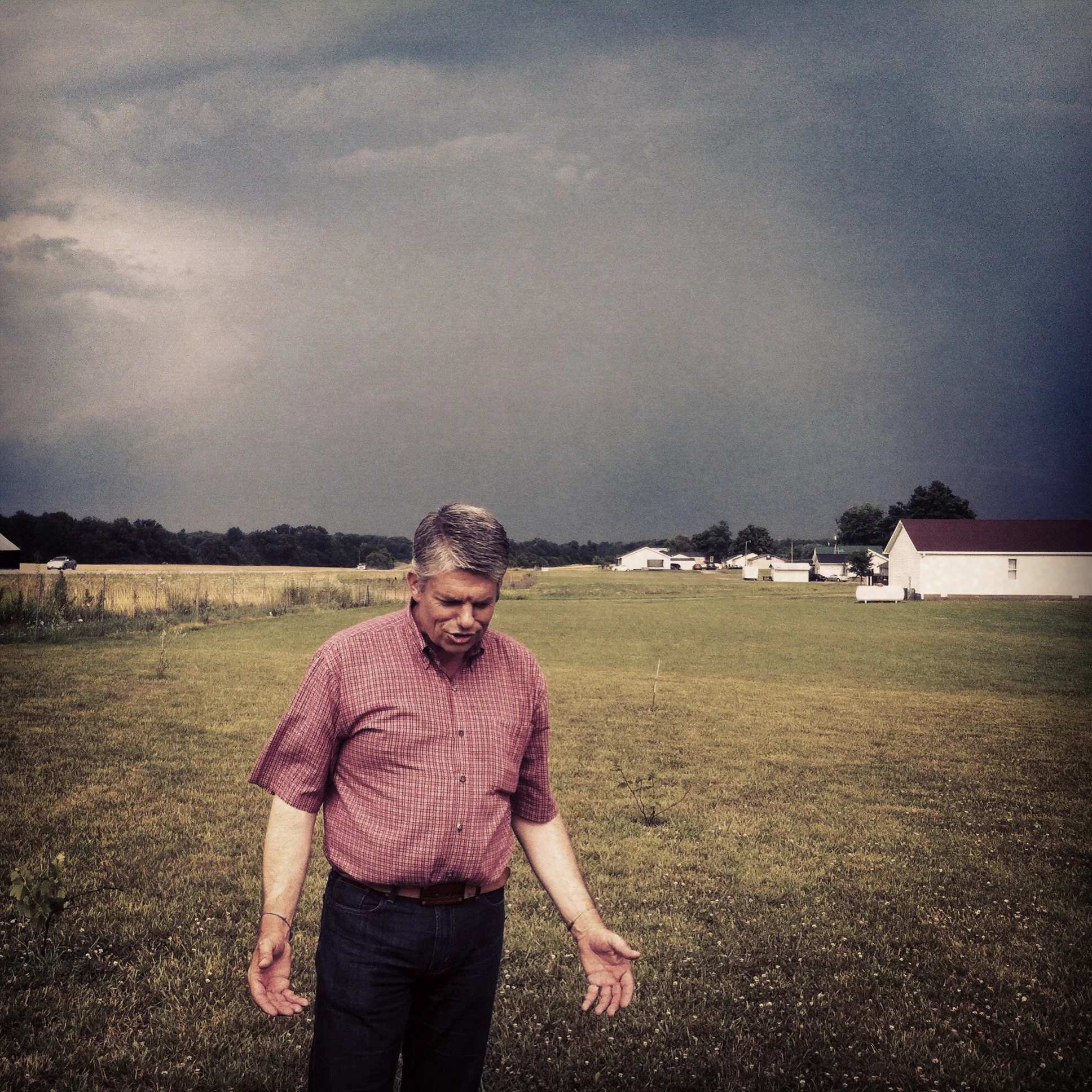 A man standing in an open grassy field with dark storm clouds overhead, several small white buildings and trees in the background.