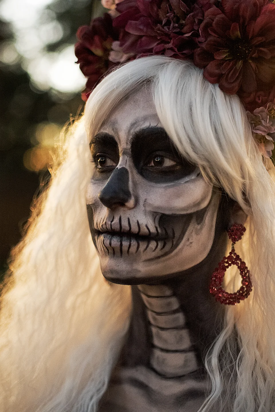 A woman with long white hair wearing traditional Día de los Muertos skeleton face paint and a floral crown with large maroon flowers, outdoor setting with bokeh background.