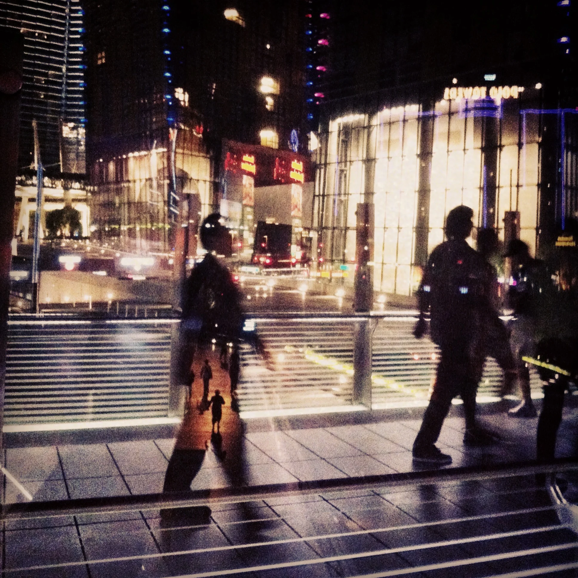Nighttime city scene with pedestrians walking past a glass building, reflecting lights and traffic.