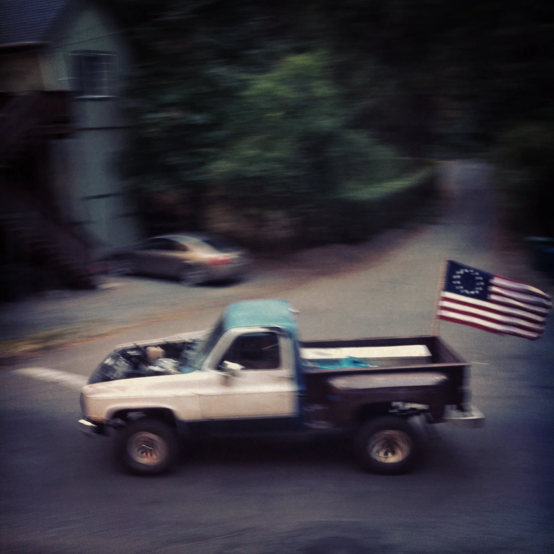 Blurry photo of a pickup truck with an American flag attached, driving on a street in front of houses and trees.