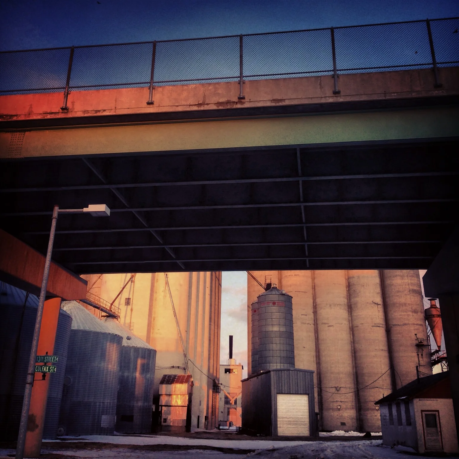 Industrial scene with large grain silos and storage buildings under a bridge at sunset.