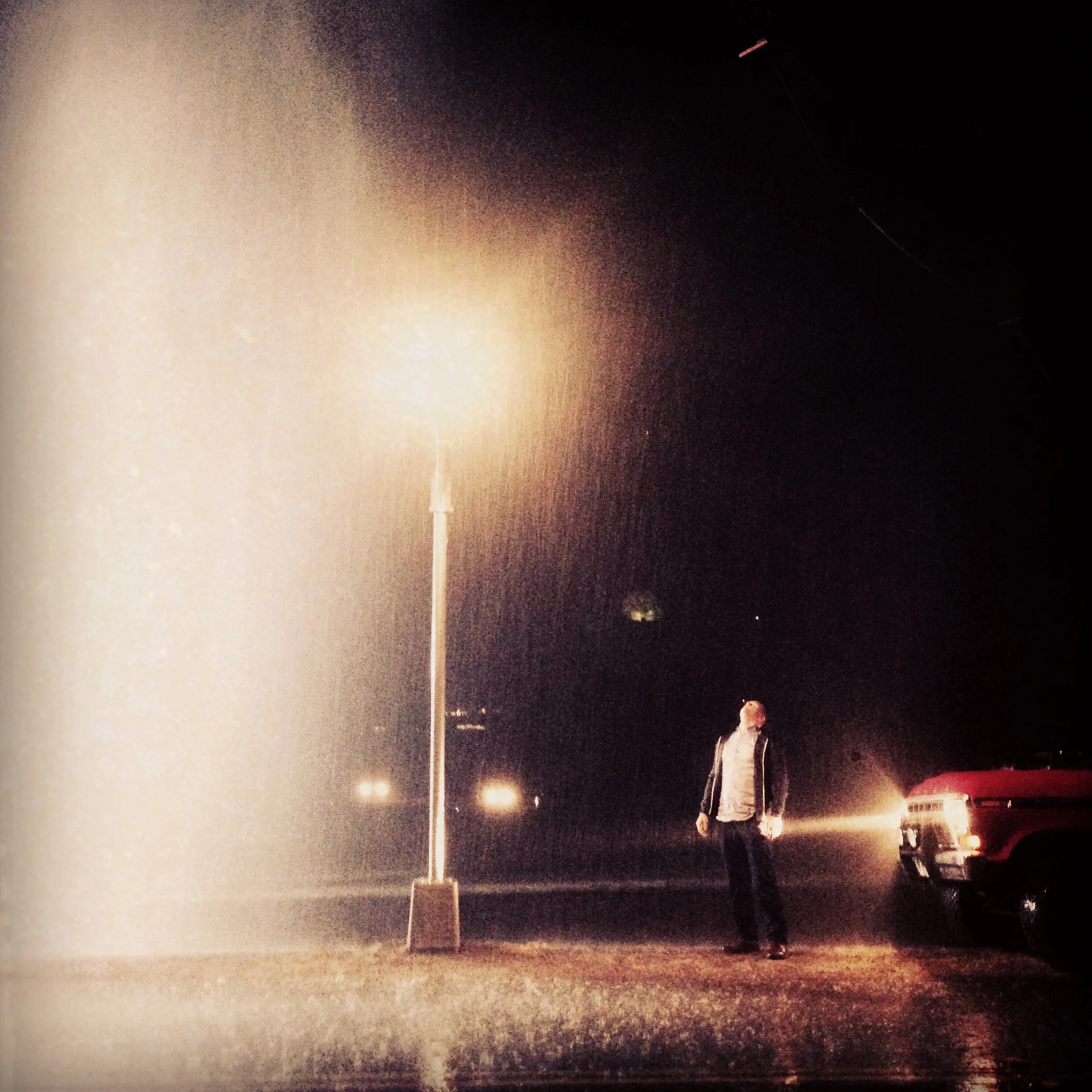 A man walking in heavy rain at night near a streetlight and a red vehicle, with visible raindrops illuminated by the lights.